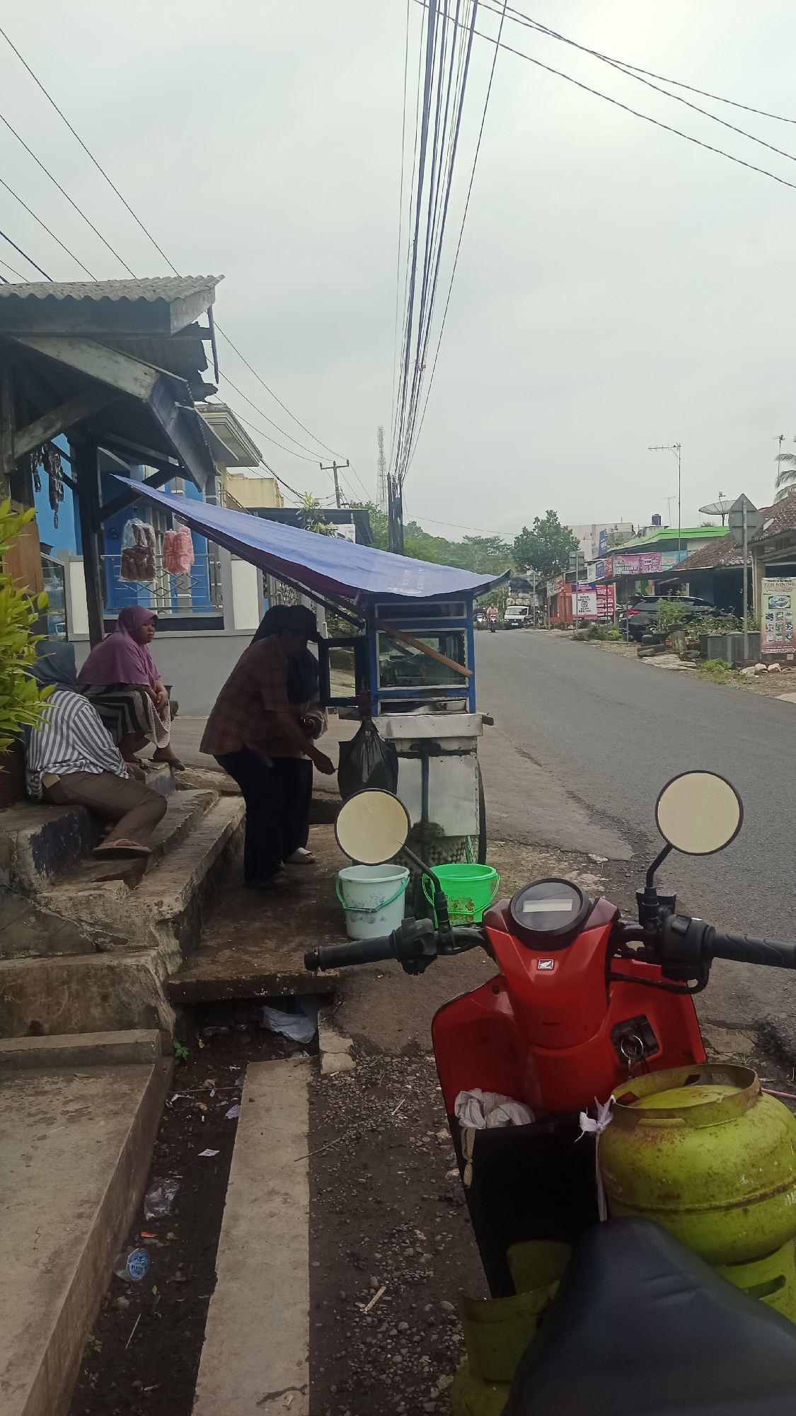 A street scene in Karyamandala, Indonesia, features a red scooter parked on the right foreground. The scooter has two green gas tanks strapped to its rear. To the left of the scooter, a food cart is set up under a blue awning. A woman in a brown patterned shirt and dark pants is standing behind the cart, seemingly serving or preparing food. Two other women are seated on concrete steps to the left of the cart. One woman wears a pink headscarf and a patterned skirt, while the other wears a striped shirt and brown pants. A blue building with some signage is visible behind them. The street runs from the foreground to the background, with buildings and vegetation lining its sides. Overhead, a dense network of power lines crisscrosses the sky. The ground is a mix of concrete, dirt, and gravel, with some debris present. The sky is overcast.