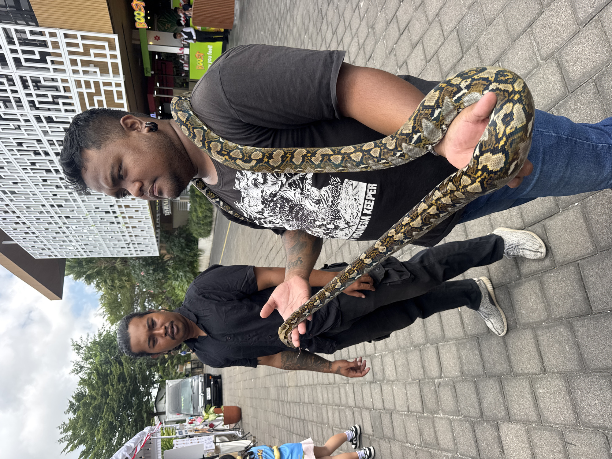 Two men are standing on a paved walkway in front of a modern building. The man in the foreground, on the right, has a large python draped around his neck and shoulders. He is wearing a black t-shirt with a graphic print and blue jeans, and is holding the snake's head in his right hand. His left arm is bent, and his hand is near his chest. The man in the background, on the left, is wearing a black shirt and black pants, and has his arms crossed. He is looking towards the man with the snake. The building behind them has a white, geometric facade and a wooden accent. A sign with "BOOST" is visible on the building. The walkway is made of grey pavers. In the background, there is a black van, some trees, and a white tent structure. The sky is overcast. The location is in Kuta Selatan, Indonesia.