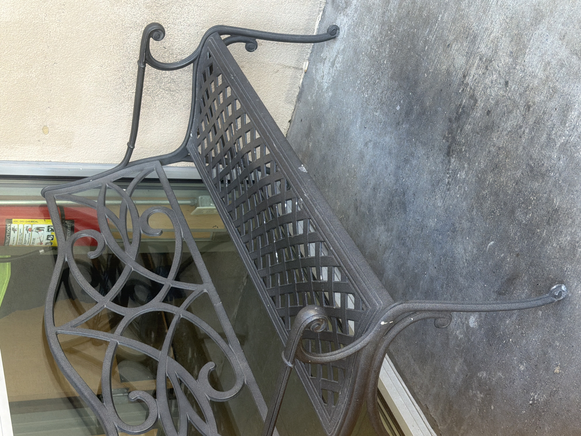 A dark metal bench with ornate scrollwork on the backrest and a lattice-patterned seat and backrest is positioned outdoors on a concrete surface. The bench is facing left, with its right armrest visible against a textured, light-colored wall. To the left of the bench, a large glass window is present, reflecting the interior of a building, which includes a glimpse of a fire extinguisher and some boxes. The concrete ground appears worn and slightly stained. The scene is static, with no individuals or discernible activity. The lighting suggests it is daytime, with shadows cast by the bench.