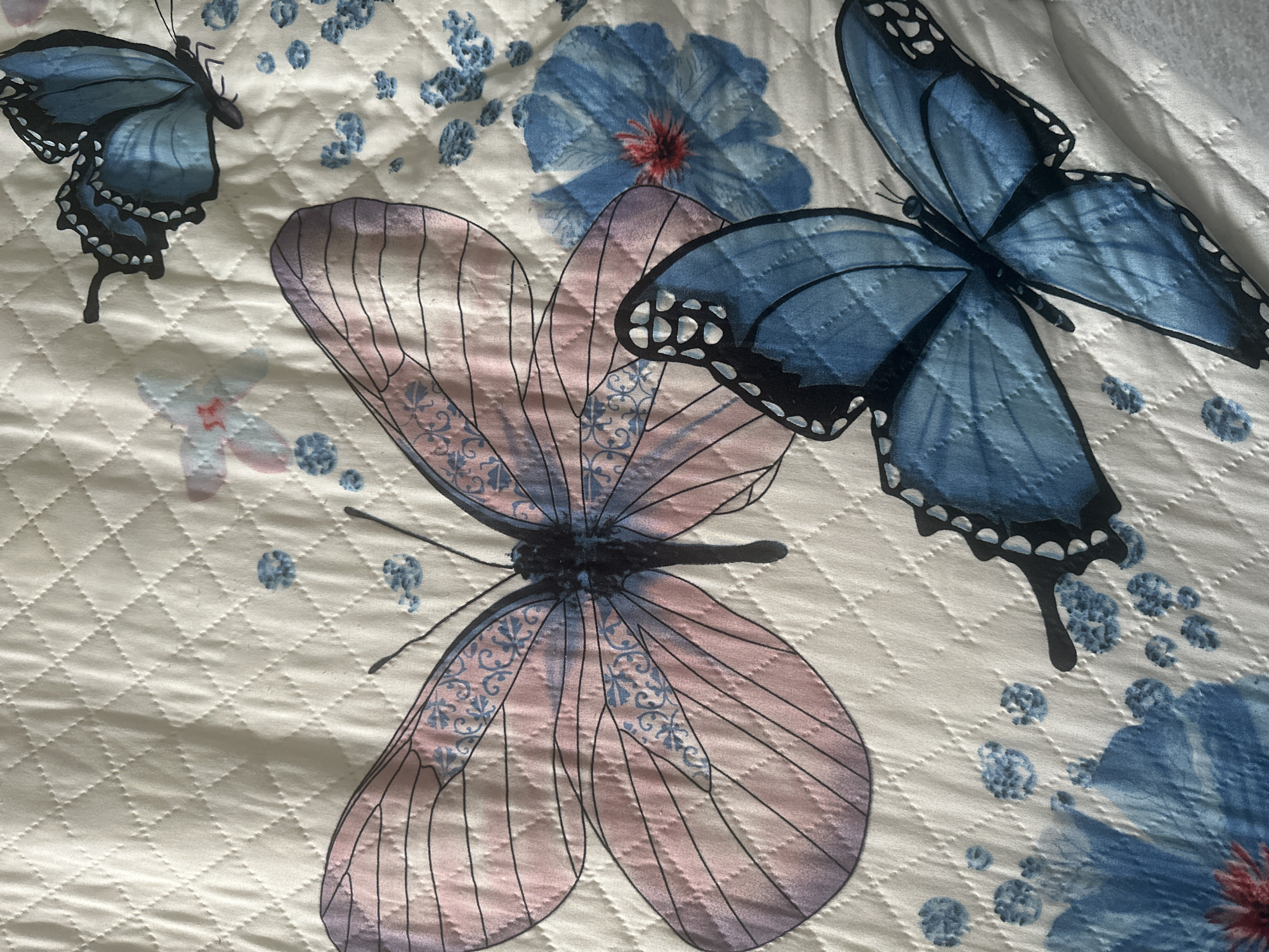 The image is a close-up of a quilted fabric with a pattern of butterflies and flowers. There are at least three prominent butterflies depicted: a large pink and purple butterfly in the center left, a medium-sized blue butterfly in the upper right, and a large blue butterfly in the lower right. Scattered around the butterflies are small blue floral clusters and larger blue flowers with red centers. The fabric itself has a diamond-stitched quilted texture. The image does not provide any information about the city of Khimki, Russia, or any other location. There are no people or actions depicted.