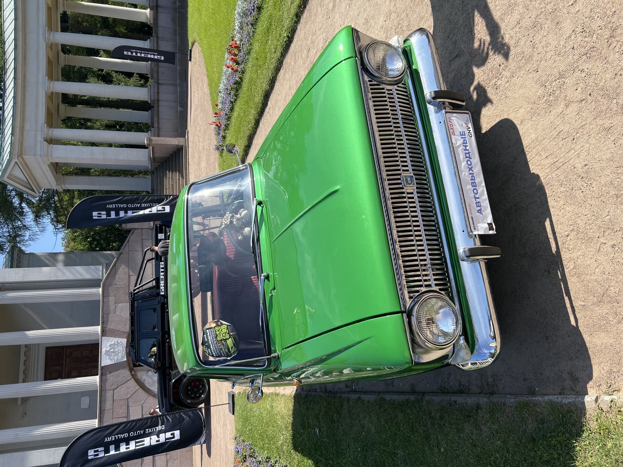 In an outdoor setting, a vintage green sedan with a shiny paint finish is parked on a gravel pathway. The car features a chrome grille, round headlights, and a chrome bumper. A license plate with Russian text and "auto.ru" is affixed to the front bumper. Inside the car, a stuffed leopard toy is visible on the dashboard. To the left of the sedan, a black pickup truck with a roof rack is parked. Two black flags with white lettering, reading "GREATS DELUXE AUTO GALLERY," are prominently displayed. In the background, a building with a classical architectural style, featuring large columns and a wide staircase, is visible. The area is landscaped with a grassy lawn and flowerbeds. The sky is clear and blue, indicating a sunny day.