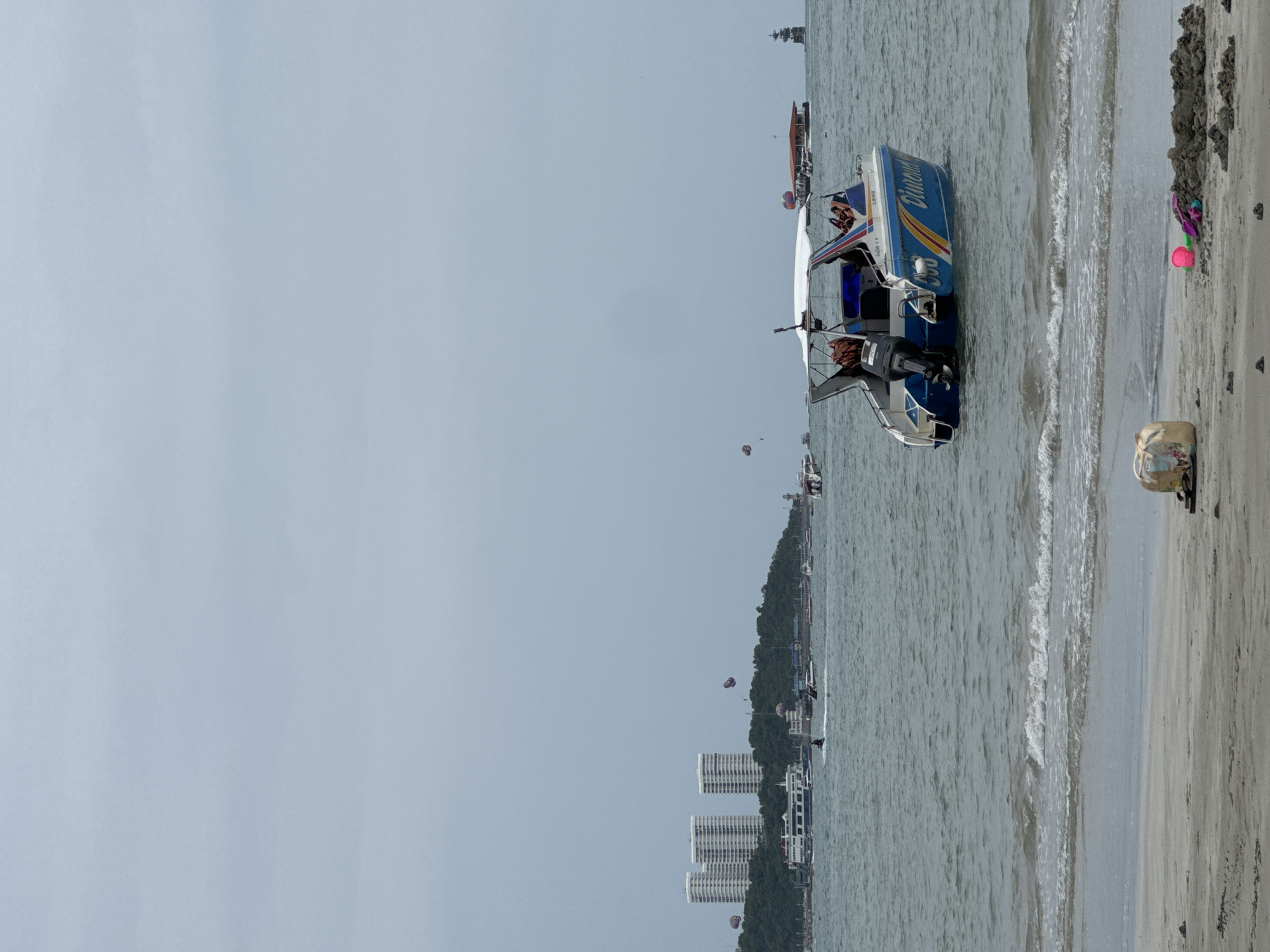 The image depicts a cloudy day at Pattaya Beach, Thailand. In the foreground, the sandy shore is visible with a beach bag and children's sand toys. The sea is choppy with gentle waves lapping at the shore. Several boats are present in the water, including a speed boat with people on board, a ferry in the distance, and other smaller vessels. In the background, the coastline features a lush green hill topped with several tall, modern buildings. Parasailing activity is evident with colorful parachutes visible in the sky over the water. The overall atmosphere suggests a typical day at a popular tourist beach.