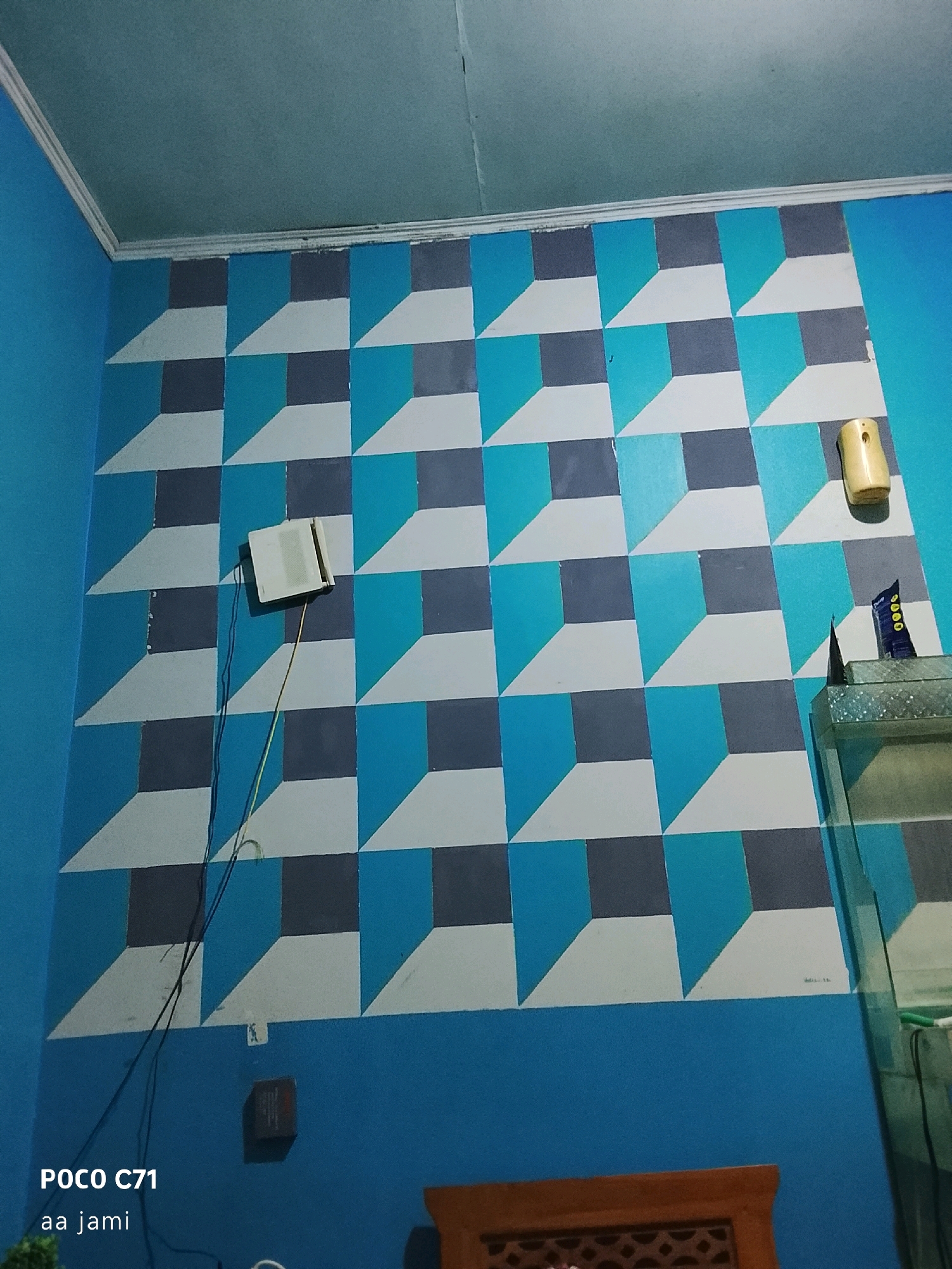 The image captures a brightly lit room with a wall featuring a visually striking geometric pattern. The pattern, composed of squares in varying shades of blue, grey, and white, creates an illusion of depth and three-dimensionality, resembling cubes or optical art. In the upper portion of the image, a white ceiling with a decorative molding is visible, along with a light blue wall on the left.
Several objects are present on the patterned wall. A rectangular white device with some visible text, possibly a router or a small speaker, is mounted on the wall and connected by a thin wire that snakes down. Above and to the right of this device, a small, light-yellow air freshener is attached to the wall. Further to the right, a corner of a glass aquarium or shelf unit is visible, with a dark blue tube or bottle resting on it.
The lower part of the image shows a solid blue wall. A small, dark-colored electrical box or outlet is mounted on this wall. At the very bottom, a portion of a wooden piece of furniture with an intricate carved pattern is visible. The room appears to be a residential interior. The text "POCO C71 aa jami" in the bottom left corner is a watermark indicating the device used to capture the image and possibly the user's name or identifier. The image was taken in Tasikmalaya, Indonesia.