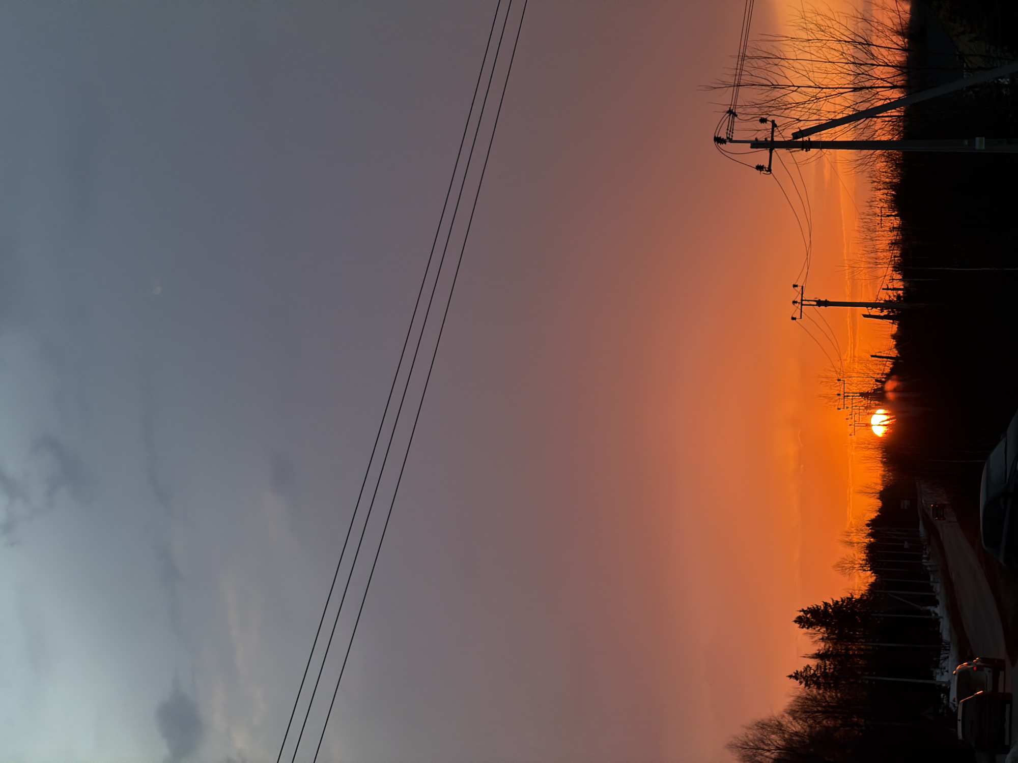 The image shows a sunset view in Izhevsk, Russia. The sky transitions from a gray-blue on the left to a fiery orange-red on the right where the sun is setting. Three power lines stretch across the left side of the frame. On the right side, silhouettes of trees, shrubs, and power poles line a road. The sun is partially obscured by the trees, casting a bright glow. A few cars are parked along the road. The scene appears to be captured at dusk.