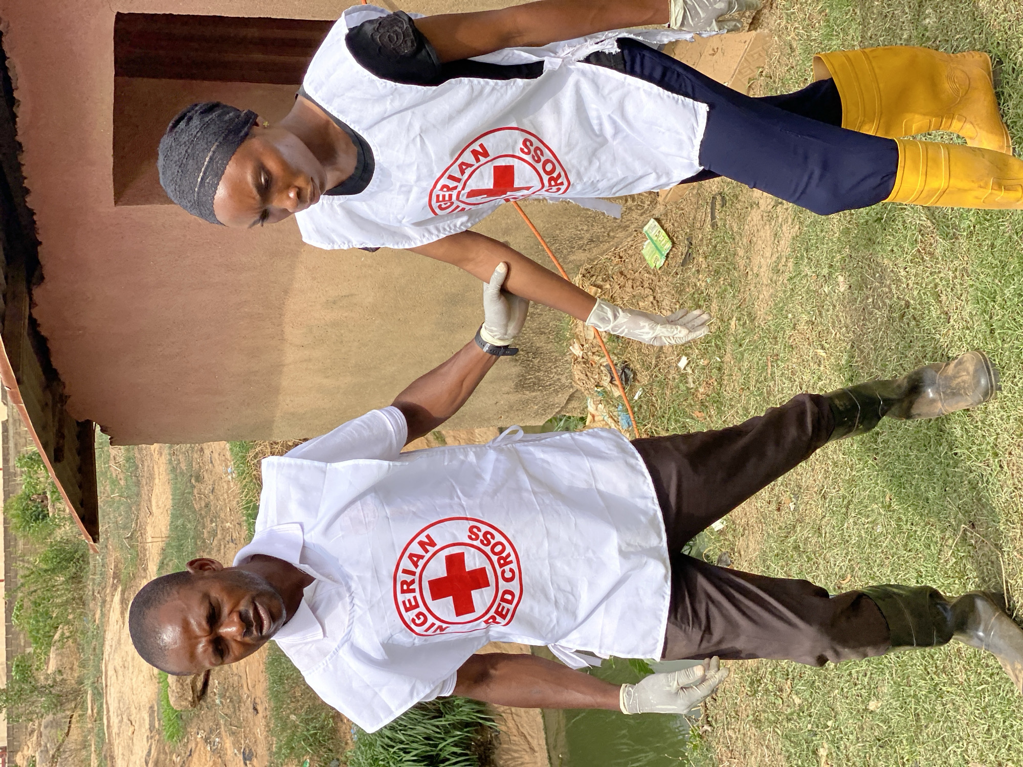 In Nassarawa, Nigeria, two individuals wearing white vests with the "Nigerian Red Cross" emblem provide medical attention. One individual assesses the arm of the other. Both are wearing latex gloves. The man wears black rubber boots while the woman wears yellow rubber boots. They stand on grass. In the background, a partially visible building stands to the left and additional greenery is visible in the distance.