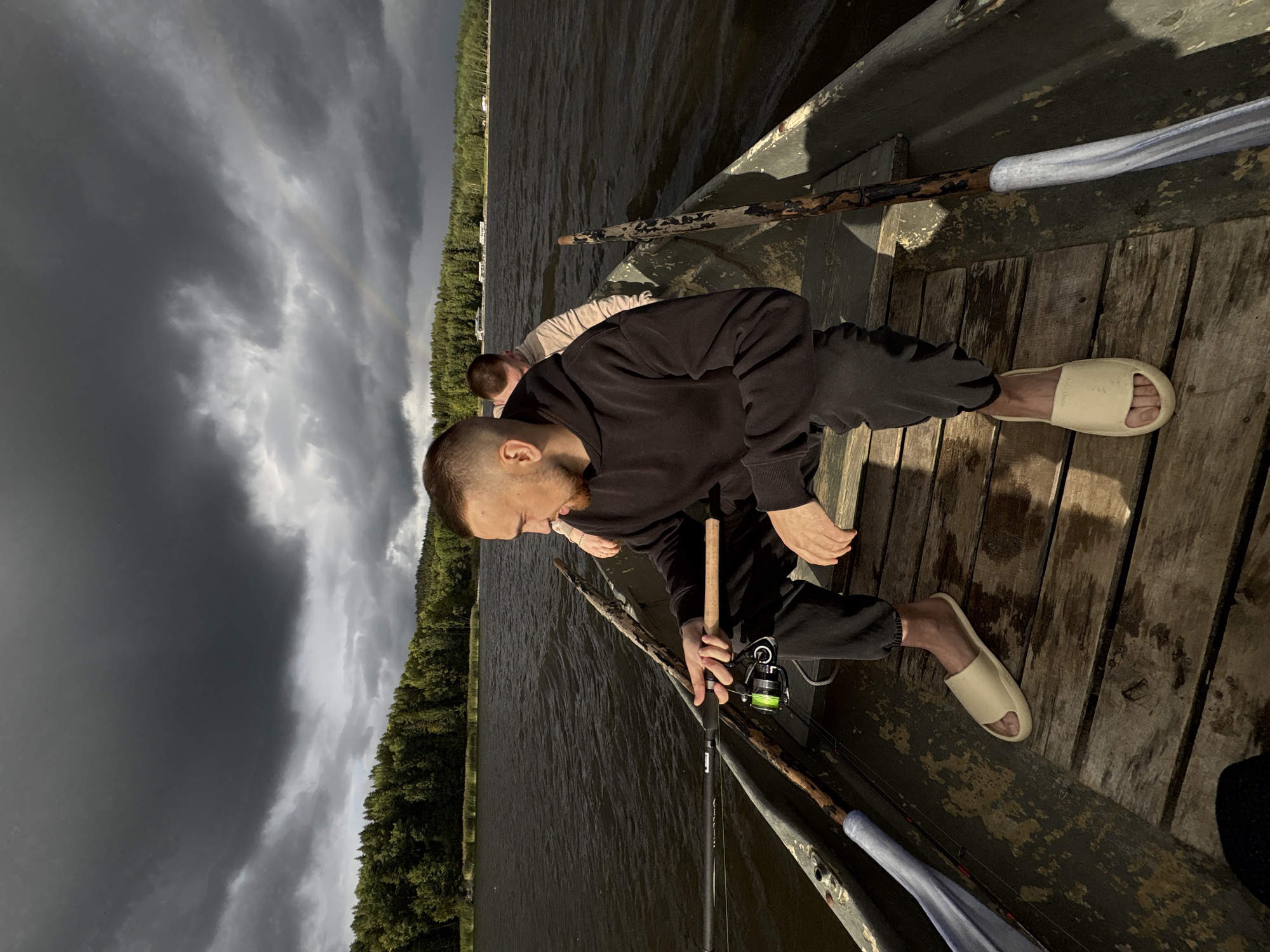In Rautalakhti, Russia, two individuals are seated in a small, weathered wooden boat on a dark, rippling body of water. The foreground is dominated by a man in a dark hooded sweatshirt and black pants, wearing cream-colored slides, holding a fishing rod with a neon green reel. His gaze is directed away from the camera towards the right. Behind him, another person, partially obscured, is seated in the boat. The boat is equipped with two oars, one on each side, with the left oar wrapped in a blue cloth. The background features a dense, green forest line bordering the water, under a dramatic sky filled with dark, heavy clouds. A faint rainbow is visible in the distance, arching across the sky. The overall atmosphere suggests an overcast, possibly stormy, day.