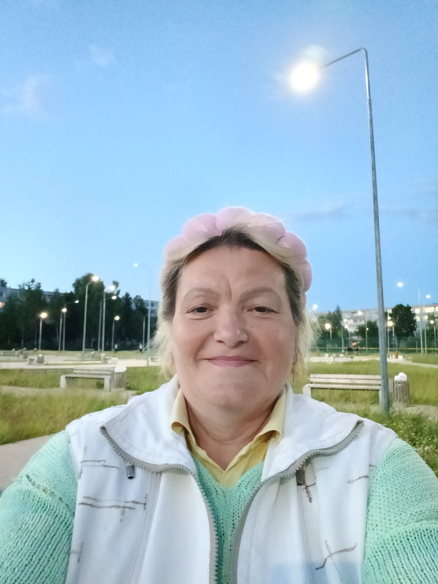 A selfie taken outdoors in the city of Sudogda, Russia, features a woman with light-colored hair and a pink headband smiling at the camera. She is wearing a light yellow collared shirt, a light green knitted sweater, and a white zippered vest over the sweater. The background shows a park-like setting with benches, trees, streetlights, and buildings in the distance under a pale blue twilight sky.