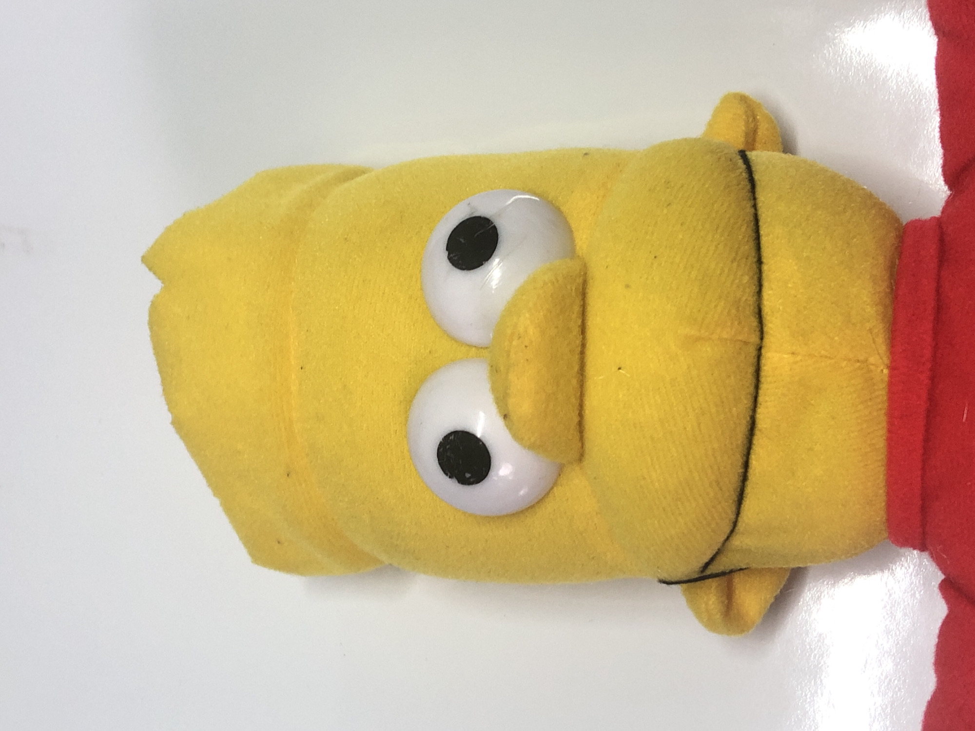A close-up, top-down shot focuses on the head of a yellow, plush toy with two large, white, cartoonish eyes, each with a black pupil. The toy has a soft, rounded shape and appears to be lying on a white, reflective surface. The toy's head is mostly yellow, with a prominent nose or snout shape protruding from the center of its face. A thin, black line extends vertically down the center of the toy's head. To the right of the yellow toy, a bright red fabric is visible, suggesting it might be part of the toy's clothing or body. The lighting is bright, creating a slight glare on the white surface and the toy's eyes. No discernible text is visible in the image, and the overall impression is of a simple, likely well-loved, stuffed toy. The scene is devoid of any contextual environmental cues, making it impossible to determine the time of day, weather, or specific location beyond the general setting of a white surface.