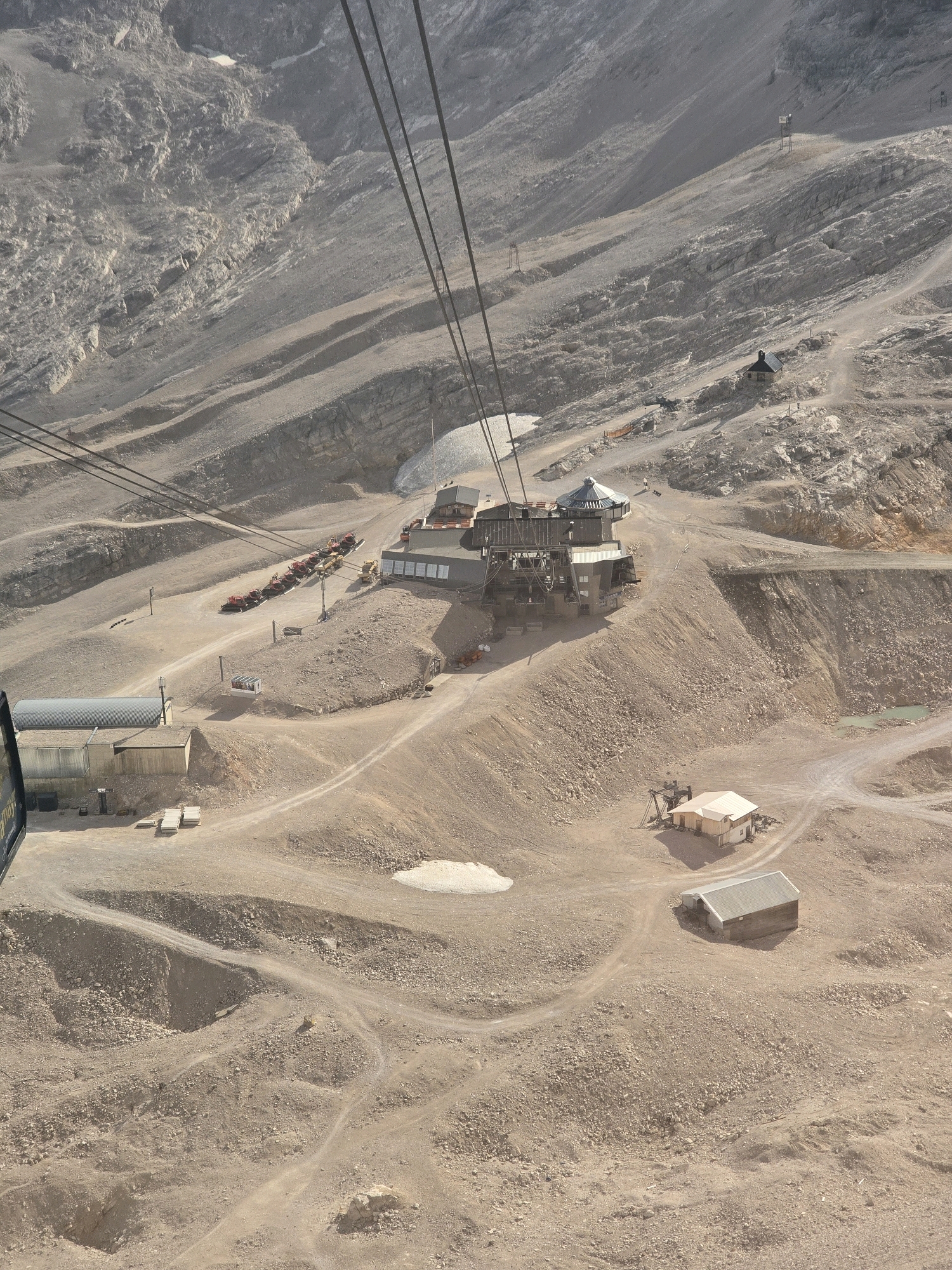 This is an aerial, wide-angle, eye-level, medium shot of a mountainous, rocky, arid landscape with a ski lift system and several buildings. The ski lift cables are prominent in the foreground, stretching diagonally across the image. Below, a large, multi-story building with a distinctive octagonal roof serves as a station for the ski lift. Various smaller structures, including garages and sheds, are scattered across the terrain, some appearing to be maintenance buildings for the ski lift or associated operations. Several red and yellow vehicles, possibly snow groomers or utility vehicles, are parked in a line near one of the buildings. The landscape itself is barren, with loose rocks and sparse vegetation. The scene appears to be at a high elevation, characteristic of a mountain resort. This location is the Zugspitze, the highest peak in Germany, situated near Garmisch-Partenkirchen.