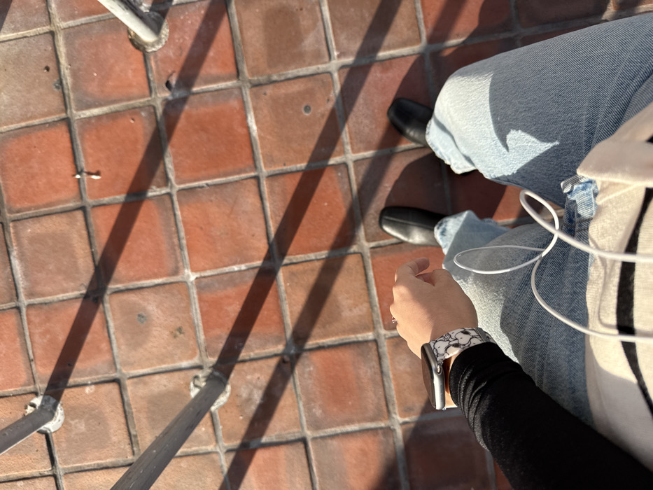 A downward, first-person perspective shot depicts a person's lower body and the tiled ground outdoors in Hialeah, United States of America. The ground consists of reddish-brown square tiles, partially illuminated by direct sunlight which casts distinct dark shadows from two metallic grey vertical poles or railings. The individual wears light blue denim jeans and black shoes. The visible left arm is clad in a black long-sleeve top, displaying a silver-colored smartwatch with a black and white Snoopy-themed band on the wrist, and a ring on the left ring finger. A white coiled wired object, consistent with earphones or a charging cable, rests on the person's lap, over a light-colored garment with horizontal dark stripes.