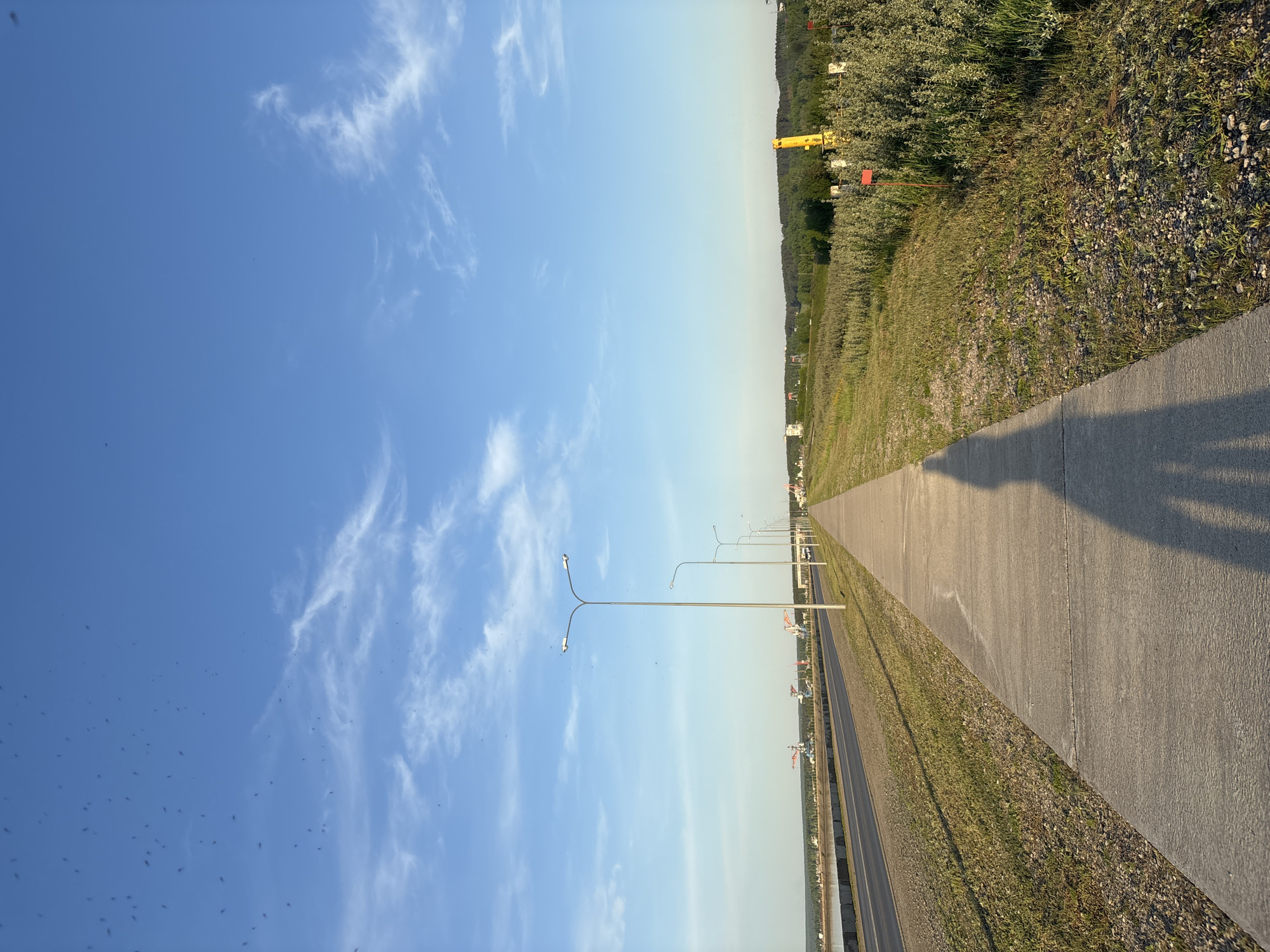 The image depicts a paved pedestrian walkway running parallel to a road in Chaikovsky, Russia. The sky is predominantly blue with scattered white clouds. Along the road, there are evenly spaced lampposts. Beyond the road, there is vegetation and possibly a river. The walkway casts a shadow, suggesting the presence of a person out of view.