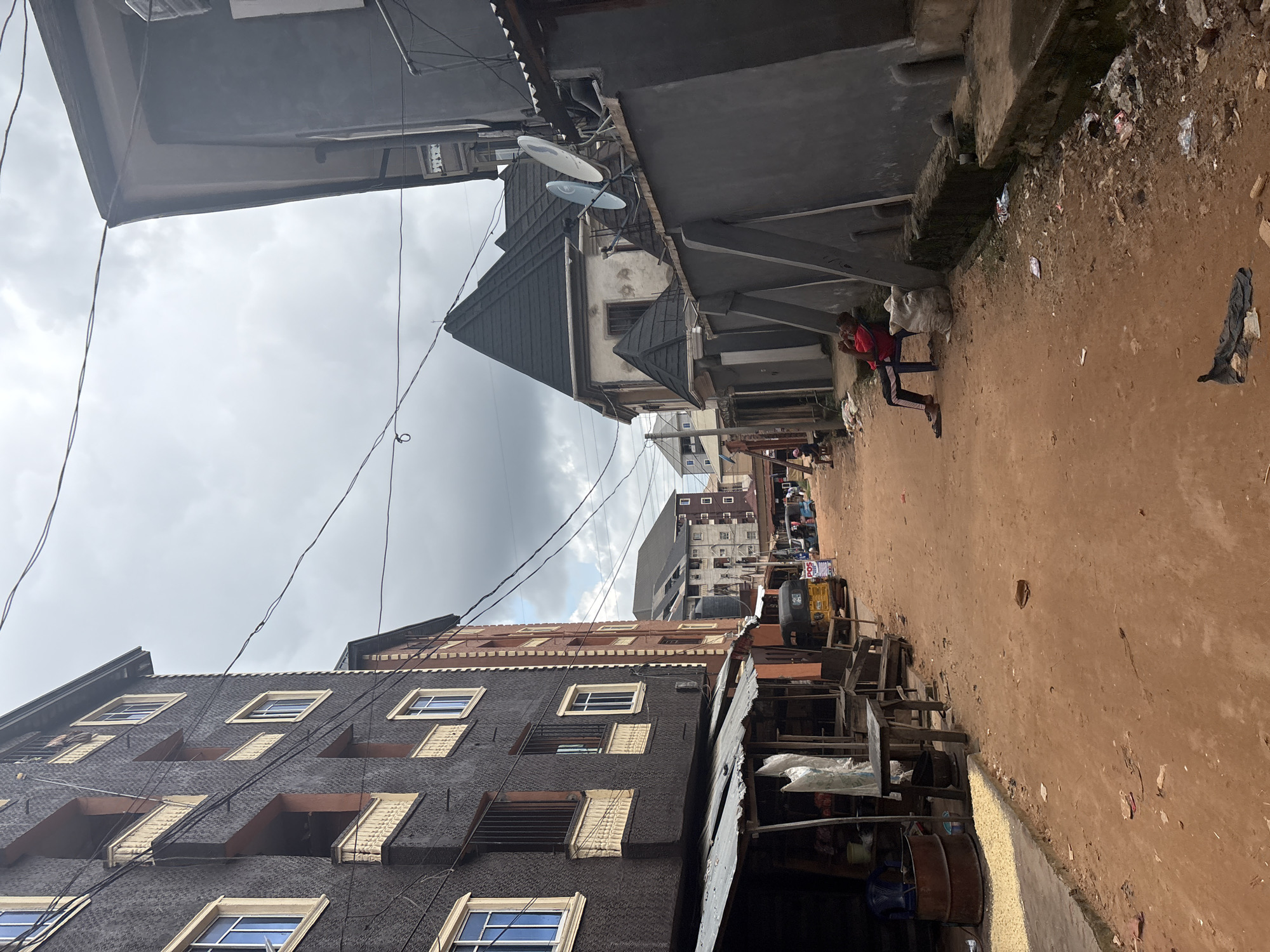 The image depicts a street scene in Nkpor, Nigeria, with buildings lining a dirt road. The buildings are multi-story structures with varying architectural styles, some appearing older and others more modern. Overhead, a network of electrical wires crisscrosses the sky, which is overcast with clouds, suggesting a cloudy day.
In the foreground, a dark-colored building with numerous windows, some with white frames and others with yellow accents, dominates the left side of the frame. Further down the street, other buildings are visible, including one with a dark, slanted roof and satellite dishes mounted on it.
The dirt road is uneven and appears to have some debris scattered on it, including trash. On the right side of the road, a few people are present. One individual in a red shirt and dark pants is seated on a chair, looking towards the camera. Other people and some vehicles, including a yellow tricycle-like vehicle, are further down the road, suggesting some activity or commerce. There are also makeshift stalls or structures made of wood and corrugated metal.
The overall impression is of a busy, somewhat cluttered urban environment. The time of day is difficult to pinpoint precisely due to the overcast sky, but it appears to be daytime. No specific emotions are readily discernible from the individuals. There is a visible sign with "POS" written on it, indicating a point-of-sale service.