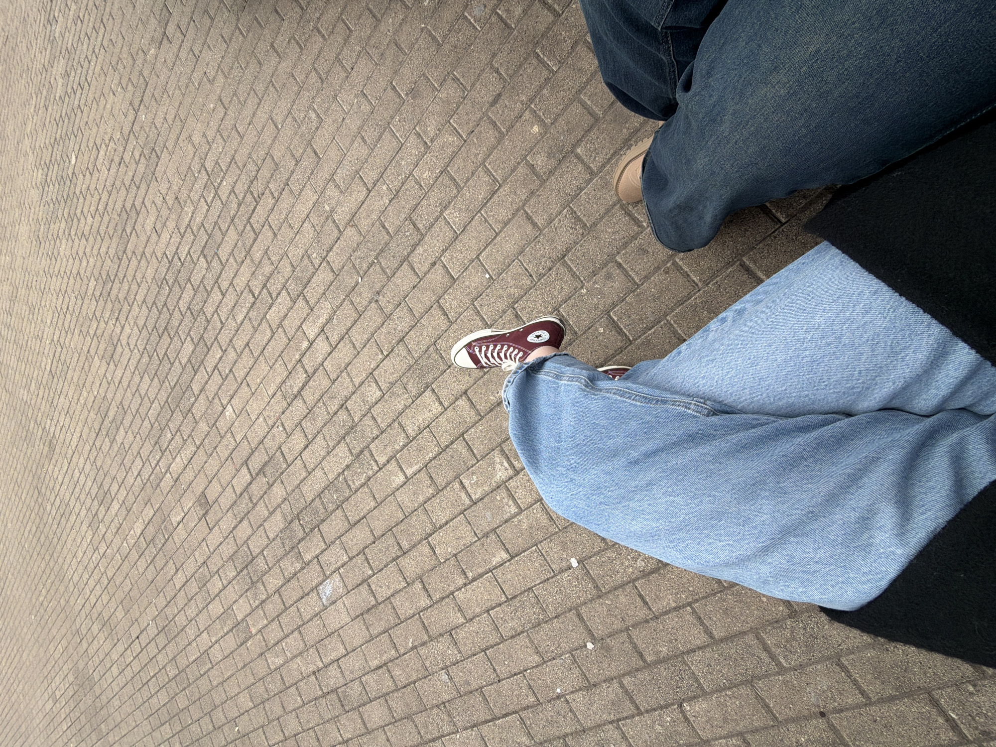 This ground-level image captures the lower bodies of two individuals on a paved outdoor surface, likely a sidewalk or public area in Chaikovsky, Russia. The ground is made of light grey rectangular pavers arranged in a diagonal, interlocking brick pattern.
In the foreground, one person wears light blue jeans and distinctive dark red (burgundy) Converse high-top sneakers. The sneakers feature white laces, white rubber toecaps, and white soles. A visible white circular patch on the inner ankle of the left shoe displays a black star and the text "CONVERSE ALL STAR." This person's legs are positioned casually, suggesting they are seated or resting.
To the right, another person is partially visible, wearing darker blue denim jeans and light brown or beige low-cut shoes. A dark black fabric, possibly a coat or bag, is visible on the far right, partially obscuring the second person's body.
The scene is illuminated by natural light, indicating daytime with clear weather conditions. No specific activity is taking place beyond the apparent moment of rest or waiting. The image focuses on the interplay of footwear, denim textures, and the patterned ground, offering a casual, everyday street-level perspective.