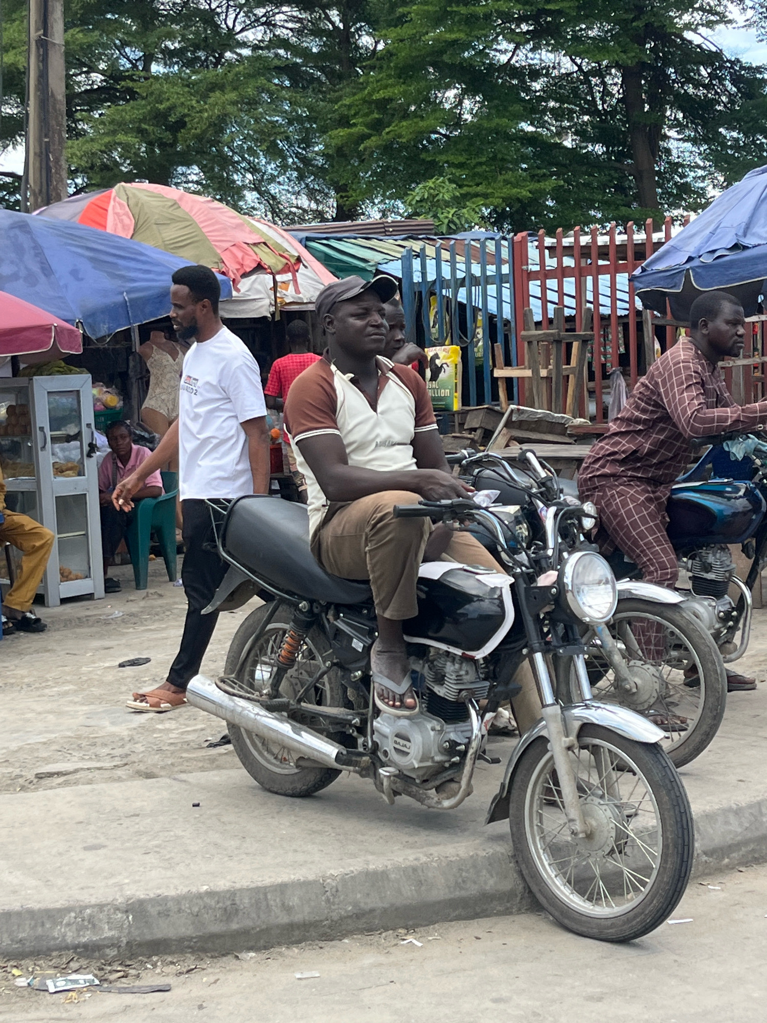 In Ado, Nigeria, an outdoor market scene unfolds under a cloudy sky, with vibrant market stalls covered by colorful awnings. In the foreground, a man wearing a brown and white polo shirt and brown pants is seated on a black motorcycle, looking to the right. Next to him, another man in a patterned brown and white shirt and pants is also on a motorcycle, facing forward. Behind them, a man in a white t-shirt and black pants stands facing left, while another individual in a red shirt is partially visible. The environment is characterized by a paved ground, a concrete curb, and assorted debris. In the background, a diverse array of market stalls are set up, displaying goods, and a wooden fence structures the area. The overall impression is one of a bustling marketplace, typical of a Nigerian city.