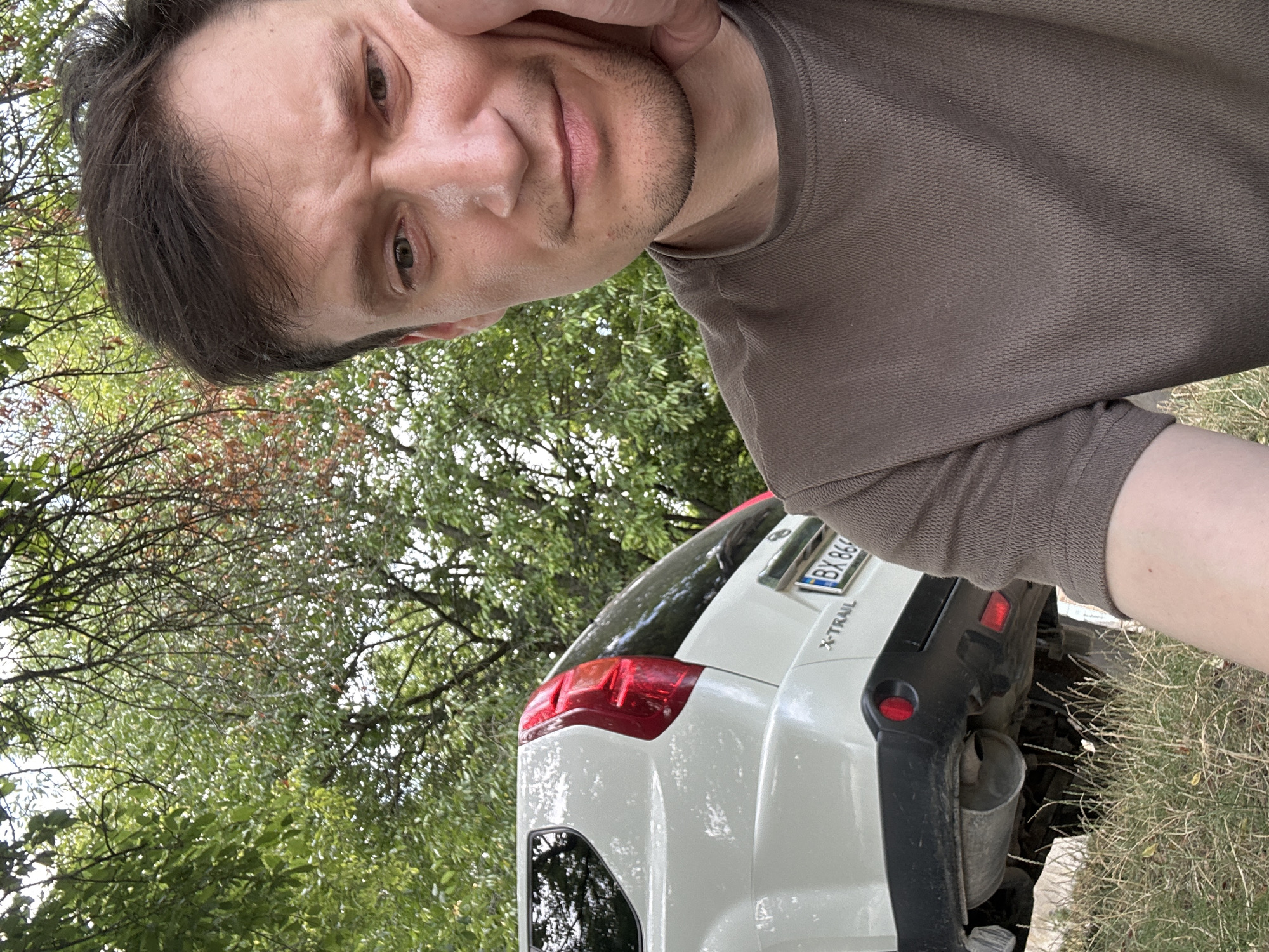 A man, who appears to be in his late 20s to early 30s, is taking a selfie outdoors. He has short, dark brown hair, light brown eyes, and a slight stubble. He is wearing a brown t-shirt. His right hand is resting on his chin, and he has a neutral expression.
Behind him, a white Nissan X-Trail SUV is partially visible. The rear of the vehicle, including the taillight, bumper, and a portion of the license plate (which reads "BX 866"), are in focus. The car is parked on a dirt or grassy area.
The background consists of trees with green and some reddish leaves, suggesting it might be autumn. The overall environment appears to be a rural or semi-rural setting, possibly on the outskirts of the city of Makiv, Ukraine.