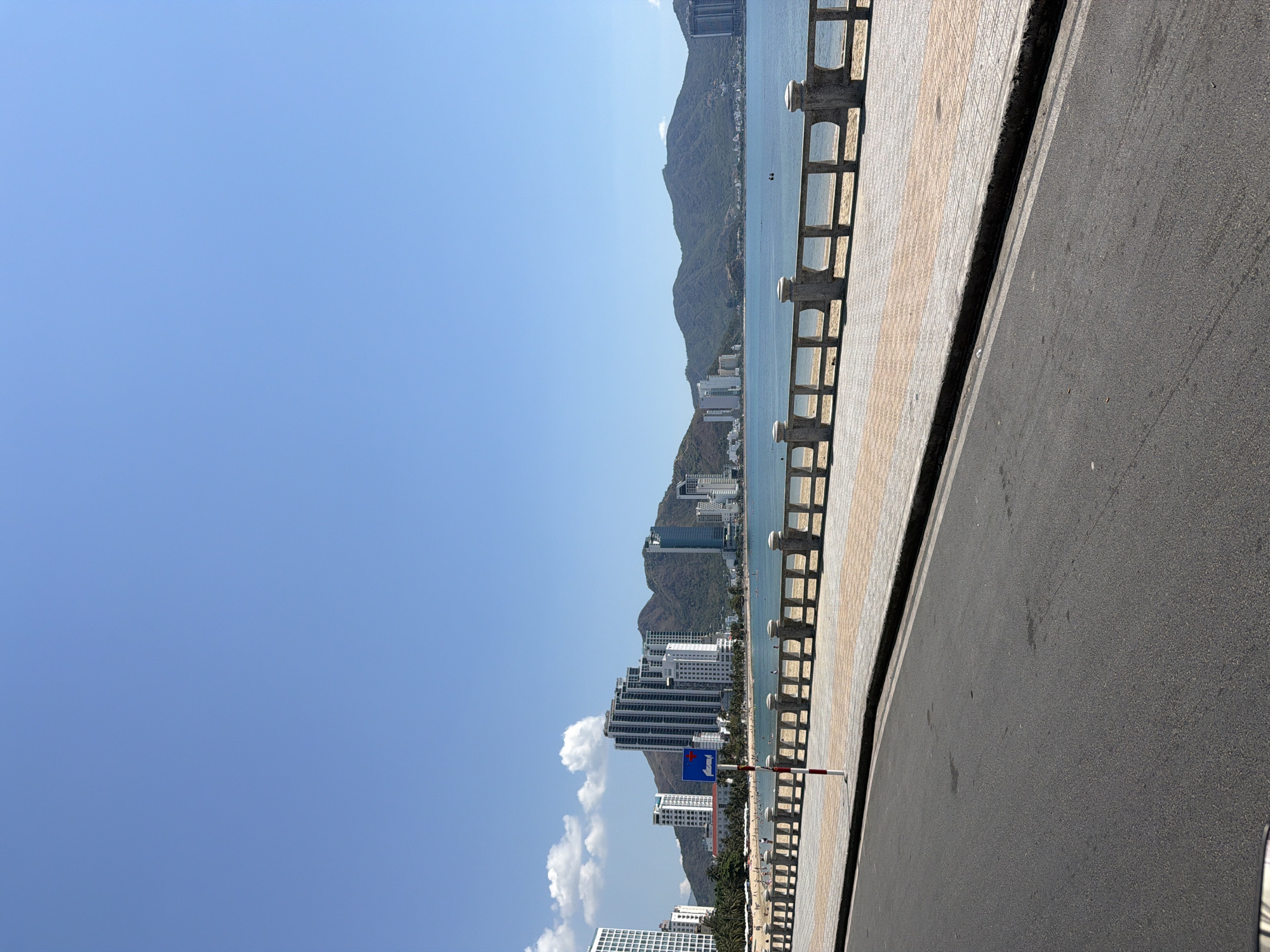 The image captures a coastal scene in Bac Nha Trang Ward, Vietnam. Concrete railings run along the edge of a tiled walkway, adjacent to a paved road. In the distance, a coastline is visible with several high-rise buildings. Behind the buildings, a mountain range rises along the coast. The sky is predominantly clear and blue.