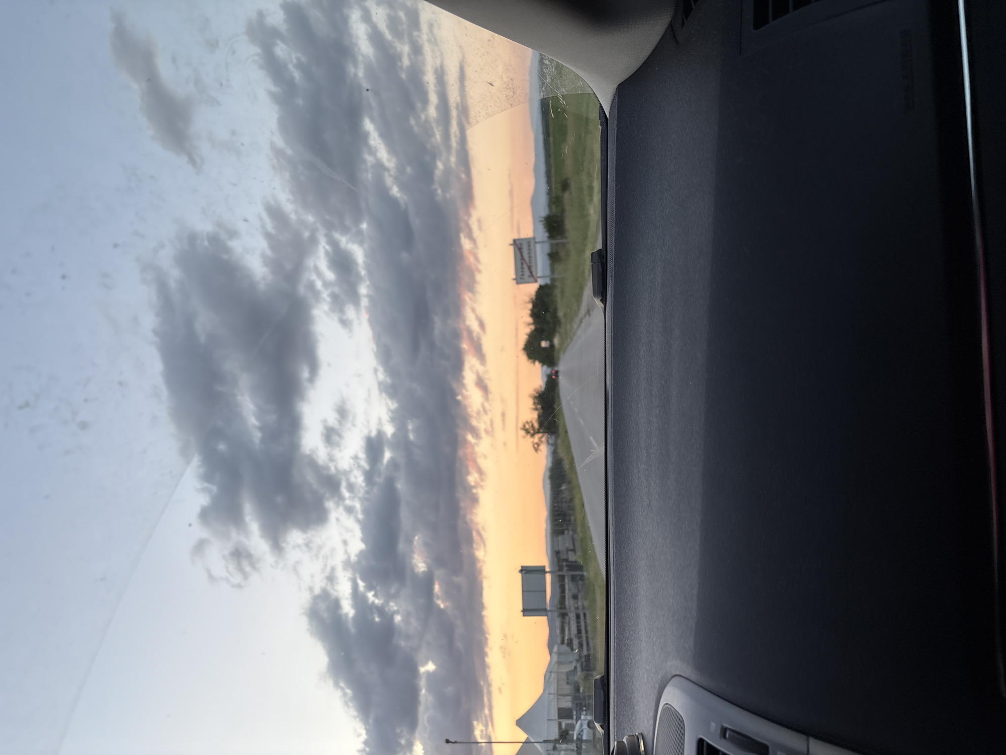 The image appears to be taken from inside a vehicle, looking out through the windshield. Outside, there is a road with signage visible, suggesting a highway or main road. The sky is filled with clouds, indicating either an overcast day or a sunset/sunrise setting with colors ranging from orange to grey. The location is likely Gelemenovo, Bulgaria, based on information provided separately. No people are directly visible in the image. The vehicle's dashboard is in the foreground, occupying a significant portion of the right side of the frame.
