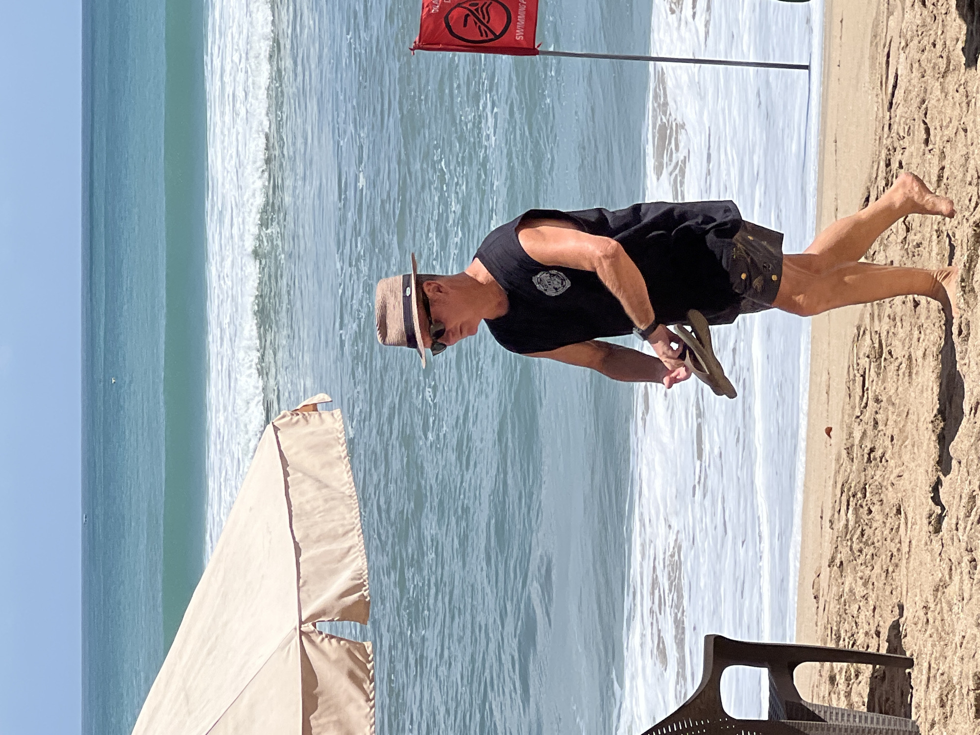 A man wearing a straw hat, sunglasses, a black sleeveless t-shirt with a circular logo, and patterned shorts walks on a sandy beach. He is holding a pair of flip-flops in his right hand. To his left, partially visible, is a brown plastic beach chair. Behind him, the ocean has waves, and a red flag with a circle and a symbol of a swimmer is planted in the sand. A light-colored beach umbrella is visible in the upper left corner. The scene is set on the beach in Kuta, Indonesia.