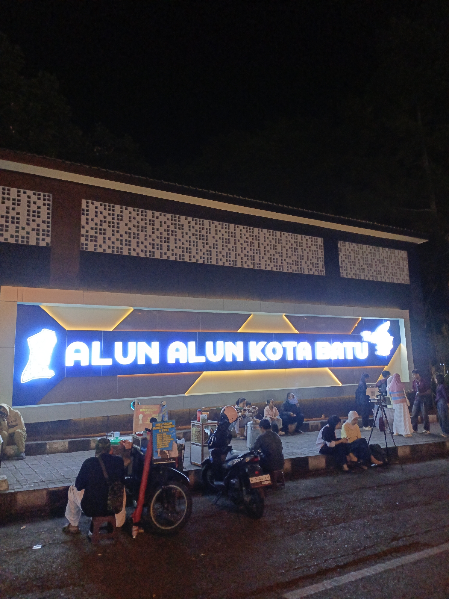 The image captures a night scene at Alun Alun Kota Batu, a public square in Batu, Indonesia. A large, brightly lit sign with blue LED lettering, reading "ALUN ALUN KOTA BATU", is prominently displayed. The building behind the sign features a dark facade with a patterned design on the upper section, composed of small, light-colored squares against a dark background.
In the foreground, several people are gathered on a tiled walkway and the adjacent street. Some are sitting on the curb, while others are seated on small stools. Various objects are scattered around, including a motorcycle, a food cart with signage, and what appears to be a tripod with a camera. The overall atmosphere suggests a casual evening gathering. The lighting is primarily from the neon sign and ambient streetlights, creating a somewhat dimly lit environment.
