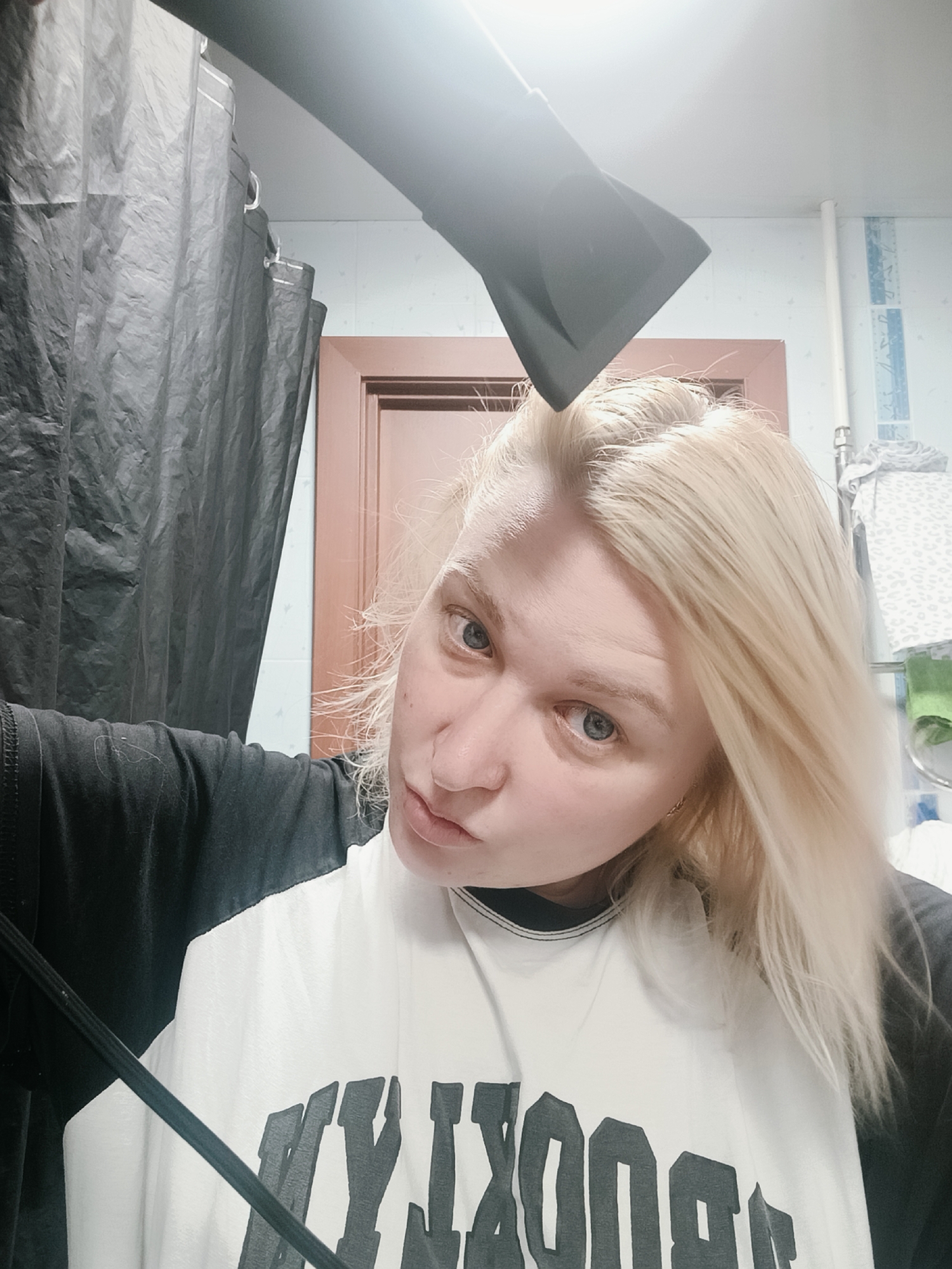 A young person with fair skin, light blonde hair, and blue-grey eyes is captured in a close-up, head-and-shoulders shot within a bathroom in Chaikovsky, Russia. They are looking directly at the camera with a casual, slightly pouting expression. Their light blonde hair appears to be in the process of being dried, as a black hairdryer is prominently visible at the top of the frame, angled towards their head.
The person is wearing a black and white baseball-style t-shirt, with the white front panel displaying dark, block-style text that reads "BROOKLYN".
The background reveals a typical bathroom setting. To the left, a dark grey or black shower curtain is pulled across. Towards the right, behind the person, a tiled wall features a vertical strip of light blue patterned tiles. A white pipe runs vertically against this wall, and a metallic towel rack holds a patterned grey towel and a green towel. A light brown wooden door is also partially visible in the background. The scene is brightly lit, likely by an overhead artificial light source, illuminating the everyday activity of hair drying.
