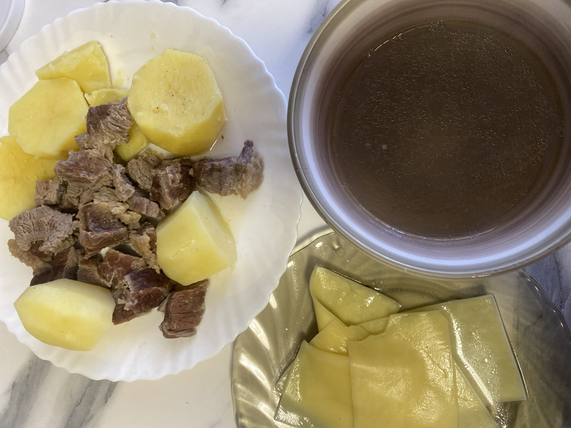 The image depicts a meal consisting of boiled potatoes and cubed beef served on a white plate, a bowl of broth, and what appears to be boiled dough squares served on a textured glass plate. These items are arranged on a light-colored countertop with a marble-like pattern. There is no indication of the location of Izhevsk, Russia, nor are there any people, actions, or interactions visible in the image.