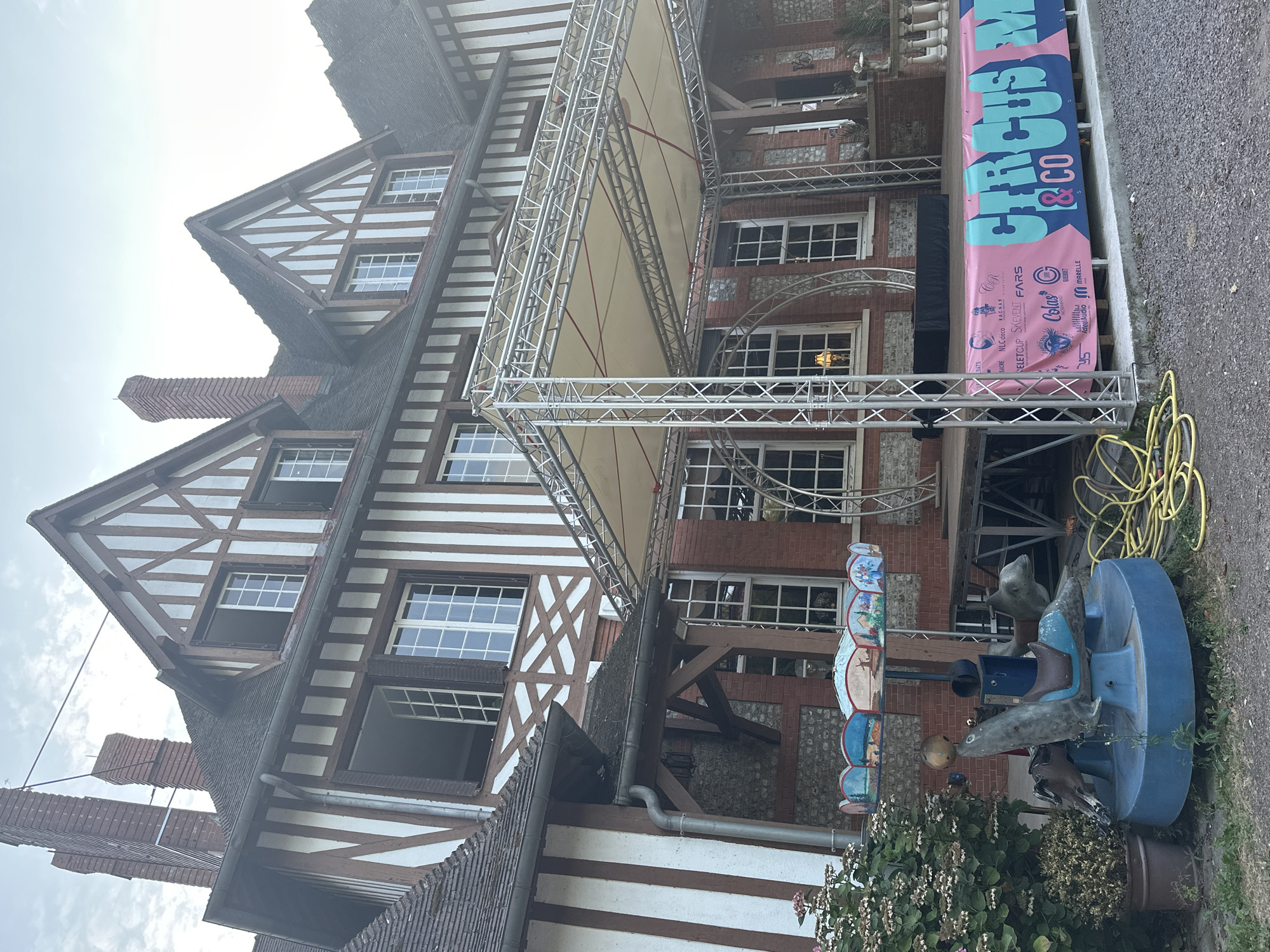 The image depicts a half-timbered building, possibly a historical residence, in Gueutteville, France. In front of the building, a temporary stage has been set up with a light-colored canopy supported by silver metal scaffolding. A pink banner with large teal lettering that reads "CIRCUS & CO" is draped across the front of the stage. Various logos are visible on the banner, suggesting sponsorships. To the left of the stage, a small, colorful carousel or merry-go-round with sea creature figures and painted scenes is present, along with a potted plant. Yellow electrical cords are coiled on the ground near the stage. The building's architecture features prominent exposed wooden beams against white plaster, with red brick sections visible. Several windows, some open, are apparent on the facade. The sky is overcast.