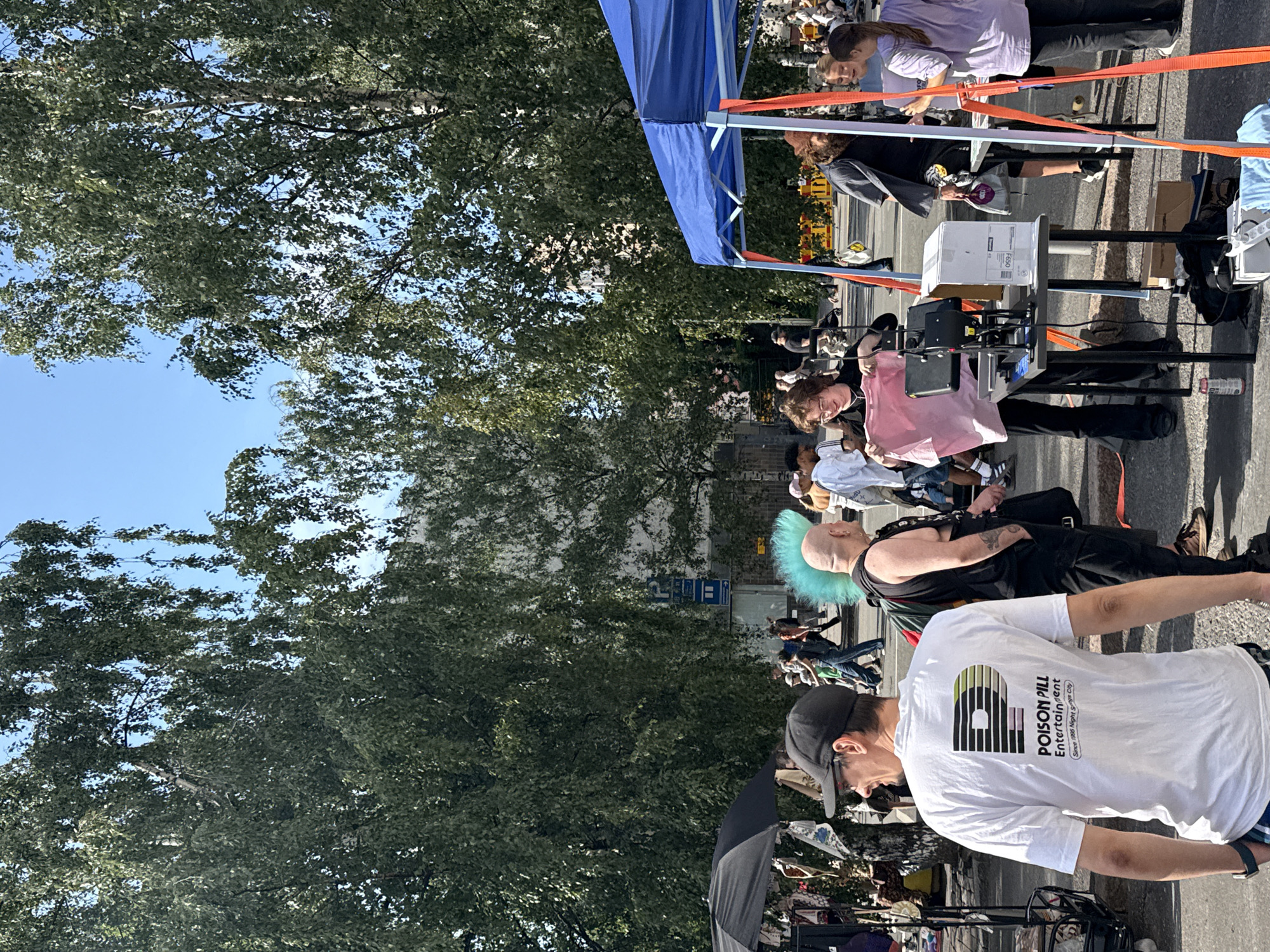The image depicts an outdoor scene on a sunny day, likely in a city setting. In the foreground, a man wearing a white t-shirt with "POISON PILL Entertainment" printed on the back is standing. Behind him, another man with a bright blue mohawk and a sleeveless black shirt is visible.
Several people are present, some interacting at tables set up outdoors. One person is holding up a pink t-shirt, seemingly for display or sale, next to a heat press machine. Another table has boxes and other items on it. A blue canopy tent is set up in the background.
The environment suggests a market or event, possibly a street fair or festival. The presence of trees and buildings indicates an urban location. The sky is clear blue, and the lighting suggests it is daytime.
While not explicitly stated, this scene is from the Puistokatu market in Helsinki, Finland.