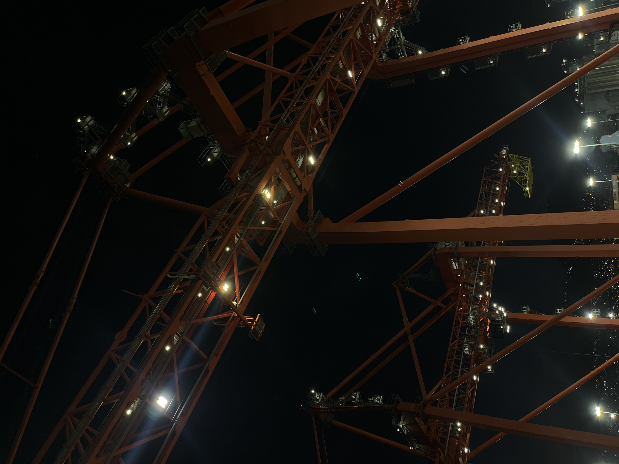 The image depicts a low-angle view of orange-colored cranes at night. The cranes are constructed with a lattice framework and illuminated with lights, creating a stark contrast against the dark sky. The location is unknown, but could be a port or industrial area.