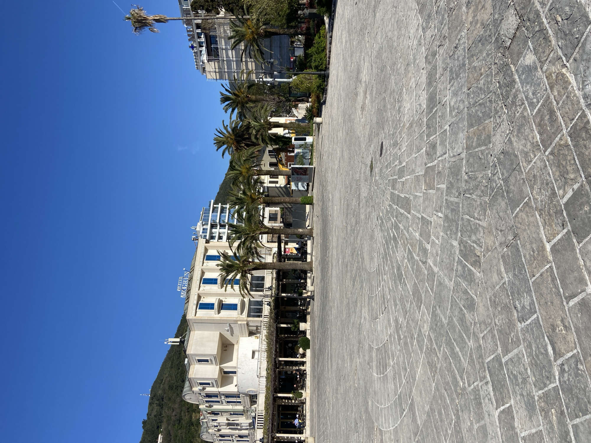 The image captures a scene in Budva, Montenegro, featuring a cobblestone promenade. On the left, the Hotel Mogren is visible, backed by a forested hillside. Palm trees line the street in front of the hotel and continue further along the road. Tables and chairs are arranged under a shaded pergola. The scene is bathed in bright sunlight with a clear blue sky. Further down the promenade are other buildings.