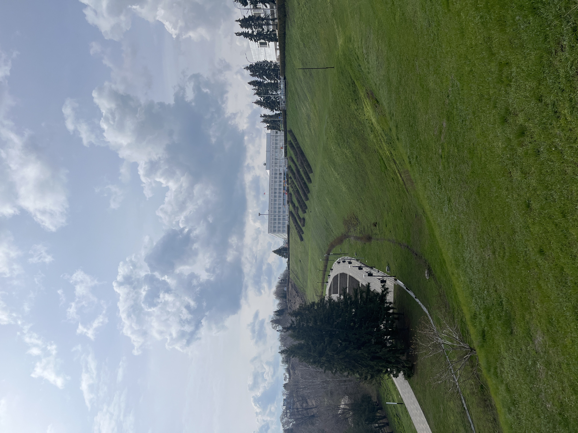 The image, correctly oriented to landscape, presents a wide-angle view of a vast, well-maintained green area under a cloudy sky. The foreground and midground are dominated by an expansive, bright green grassy slope, suggesting a park or large institutional grounds. In the upper midground and background, a large, multi-story white building with numerous windows is visible, possibly a government building or a significant institution in Ulyanovsk, Russia.
In front of the building, a series of parallel, neatly trimmed dark hedges or bushes line the landscape. Tall evergreen trees, resembling pines or firs, stand along the ridge near the building and further down the slope. A paved pathway, light in color, curves through the lower left portion of the scene, bordered by additional trees, some of which are deciduous and appear bare, hinting at early spring or late autumn. Modern lampposts are spaced along this pathway. A distinct white, arched structure, possibly a covered entrance or shelter, is located near the path.
The sky is partly cloudy, featuring both light and darker grey cumulus clouds, indicating an overcast but bright day. The diffused lighting suggests it is late morning or early afternoon. No people or specific activities are visible, contributing to a tranquil and serene atmosphere. The scene depicts a grand, public-access area, likely a park or square, connected to a prominent building. No visible text is discernible in the image.