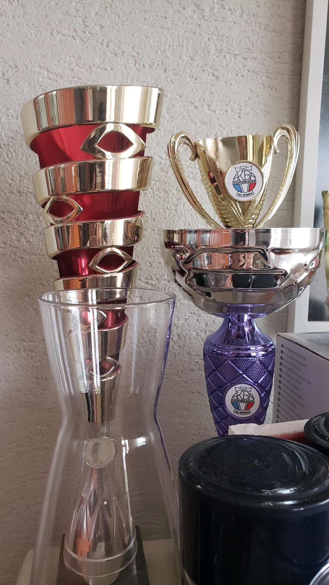 The image depicts a collection of trophies and decorative objects displayed on a shelf against a textured light-colored wall. In the foreground, a tall, clear glass vase with a metallic base is visible. To the right of the vase, a silver trophy with a purple patterned base stands prominently. This trophy features a circular emblem with the letters "AKBA" and some text below it. Behind this silver trophy, another golden trophy with similar handles and a circular emblem is partially visible. To the left of the vase, a stack of red and gold trophy components, possibly part of a larger trophy, are arranged vertically. The overall setting suggests a domestic or personal display area. There is no indication of a specific location in the city of Prešov, Slovakia, within the visual information provided.