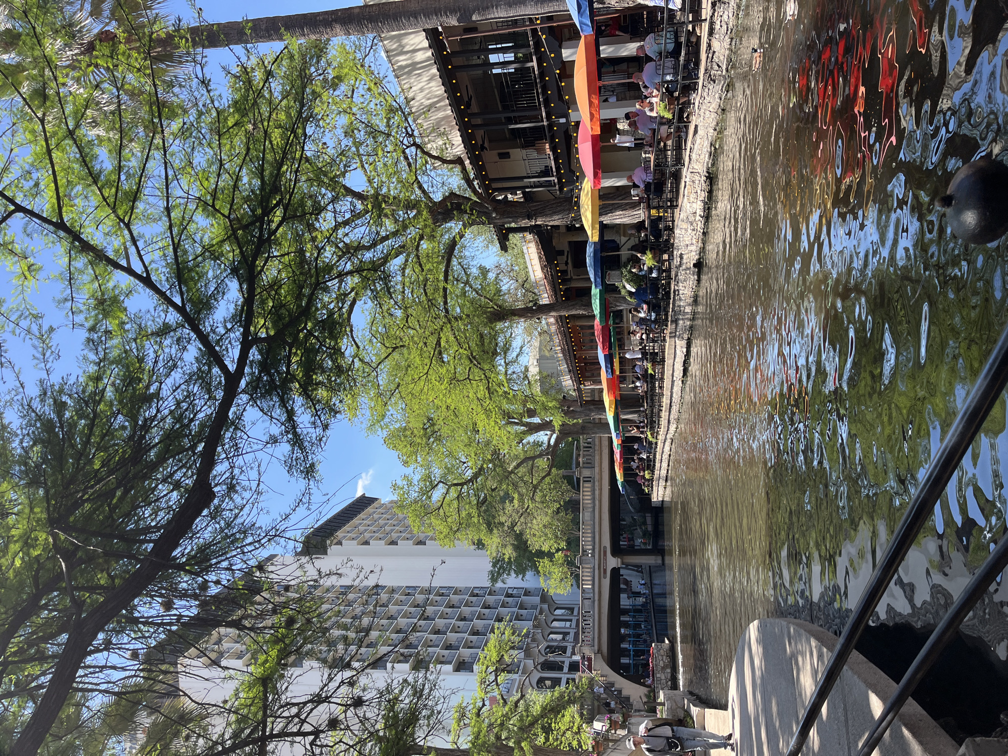 This is a photograph of the San Antonio River Walk in San Antonio, Texas, United States. The perspective is from a slightly elevated position near the water level. The river flows between buildings. Along the riverbank are restaurants with patrons seated at tables, many shaded by colorful umbrellas. A tall white building with many balconies is visible in the background. The river's surface reflects light and the surrounding environment. Trees line the riverbank. A bridge spans the river. People are walking on the walkways alongside the river. A metal railing is visible in the foreground.