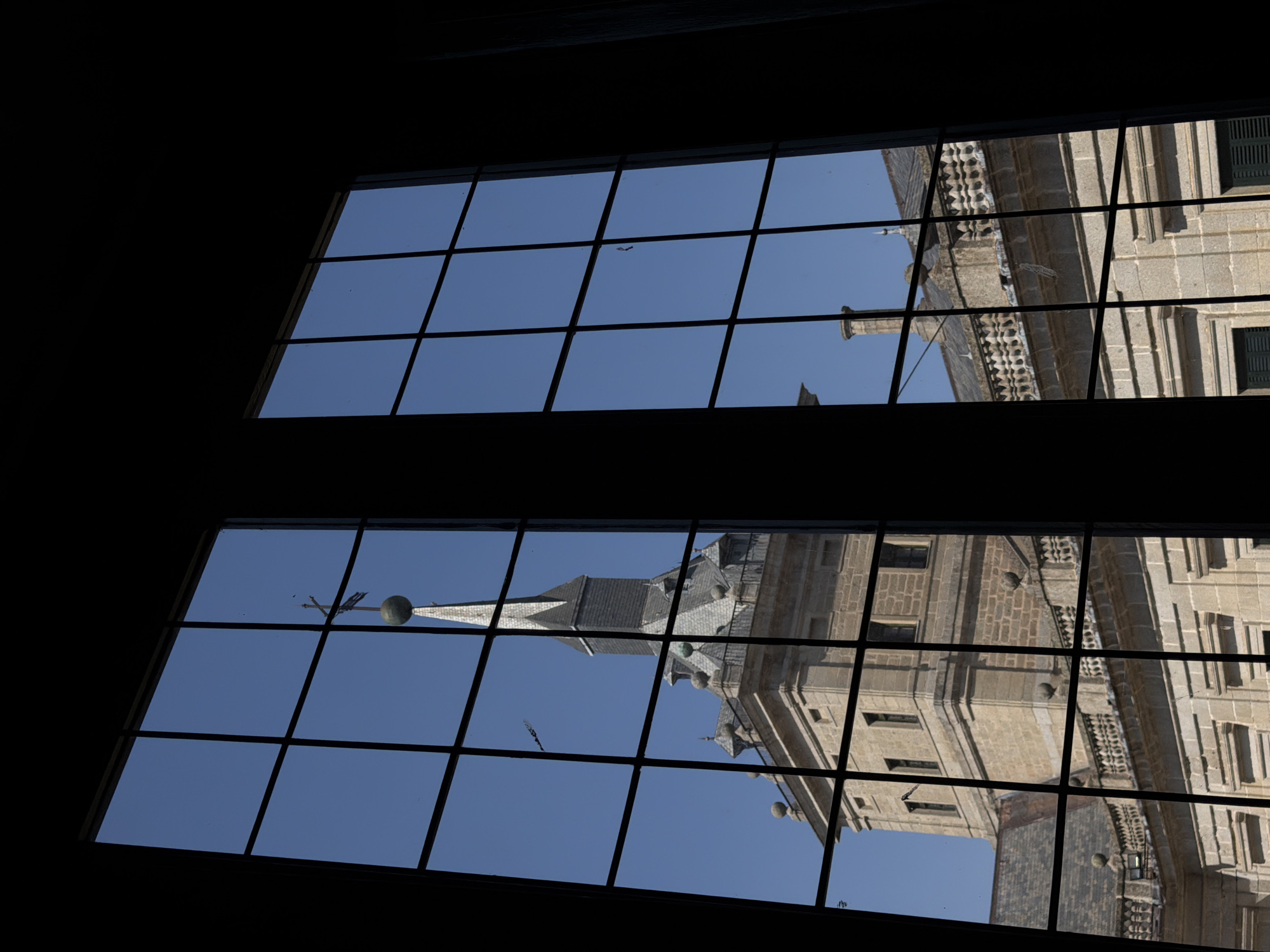 This is a medium shot of the Royal Monastery of San Lorenzo de El Escorial in San Lorenzo de El Escorial, Spain, viewed through a large, multi-paned window. The window is divided into a grid of dark mullions, with the building and sky visible through the glass. The building is constructed of light-colored stone, with a prominent tower topped by a spire and a weather vane. The architecture features classical elements, including balconies with ornate railings and arched windows. The sky is a clear, bright blue. The scene is shot from inside, with the window frame and surrounding darkness creating a strong contrast with the brightly lit exterior. There are no people visible in the image.