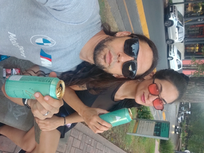 A man and a woman pose for a selfie outdoors. He wears sunglasses and a grey Motorsport t-shirt, holding an open turquoise can. She wears pink sunglasses and a black top, also holding a turquoise can. Cigarettes are near the man. A street with parked cars is visible in the background, along with a bus stop sign. The photo is taken from a slightly high angle, tilted counterclockwise.