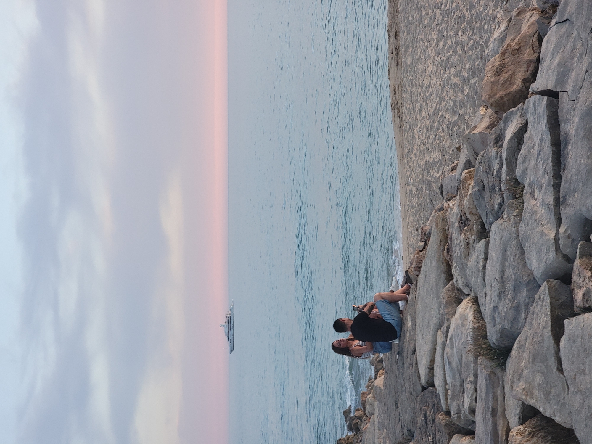 A long shot captures a serene coastal scene at dusk, with a large yacht anchored on the horizon. In the foreground, a couple is seated on a rocky embankment overlooking the water. The woman, wearing a striped top and denim shorts, is seated with her legs crossed. Beside her, a man in a black t-shirt and blue shorts holds a mobile phone, appearing to take a photo or record a video. The sky is painted with soft pastel hues of pink and blue, transitioning into the calm, cool tones of the sea. The rough texture of the rocks contrasts with the smooth expanse of the ocean and the sandy beach to the right. The scene suggests a moment of shared quietude and observation in a tranquil coastal setting, possibly along the Quarteira coastline in Portugal, given the typical presence of such yachts and the Mediterranean ambiance.