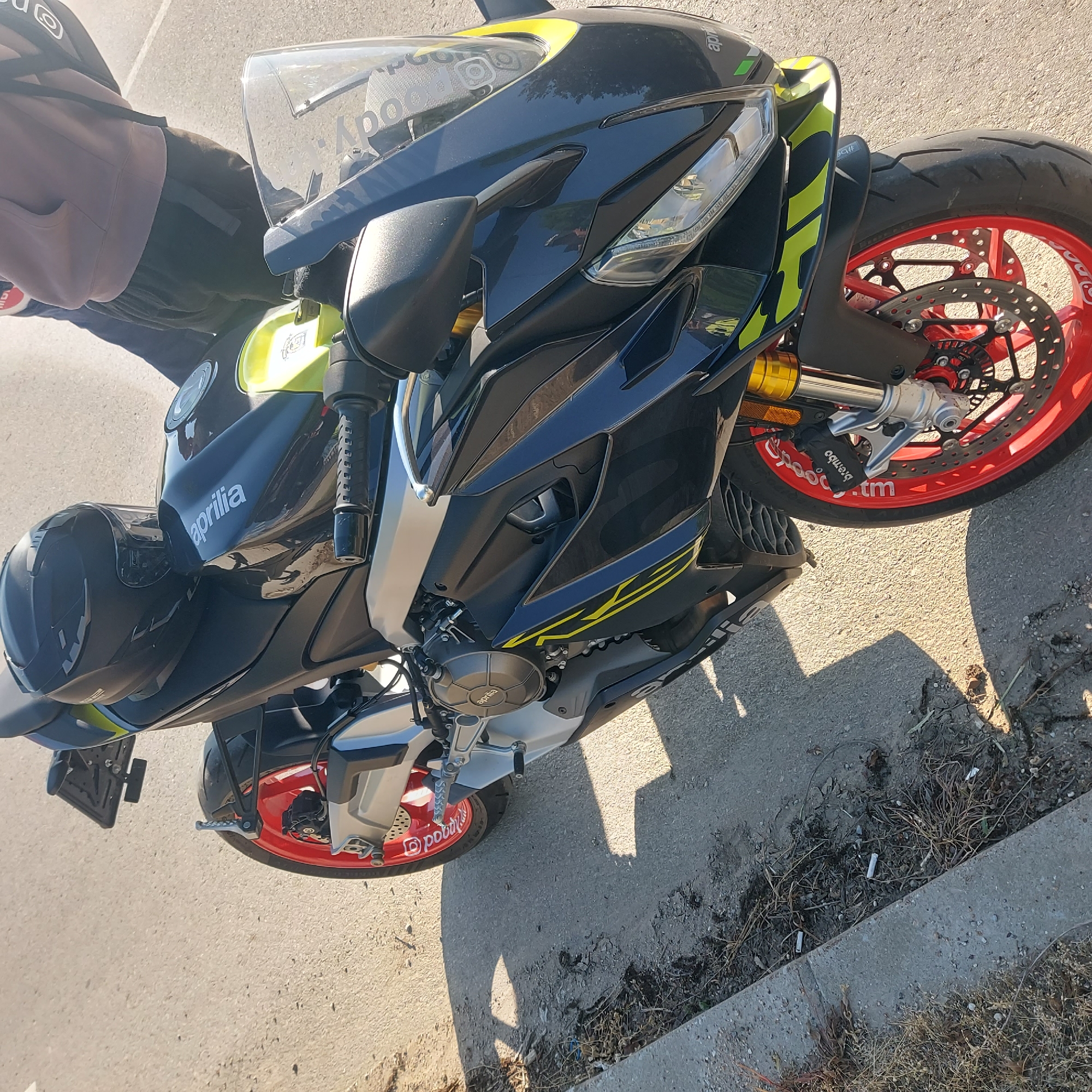 A black Aprilia motorcycle, parked on asphalt, is the primary subject. The motorcycle has red rims and yellow accents. A black helmet is on the motorcycle. The setting is a roadside with a concrete curb, and sparse vegetation in the curb area. Part of a person wearing a brown hoodie and dark trousers is visible to the left of the motorcycle. The photo was taken during the day, casting shadows on the asphalt. The image does not provide information regarding Strazhitsa, Bulgaria.