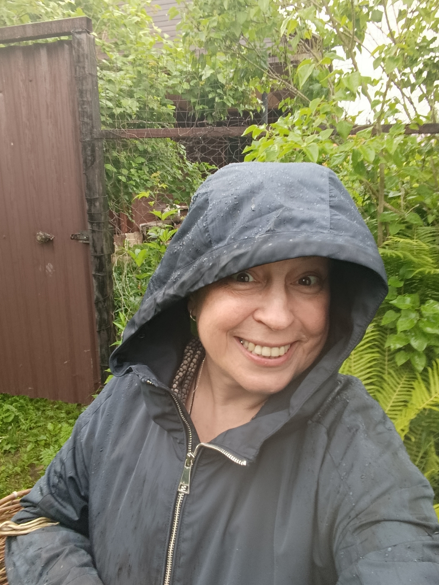 A woman in her 50s, wearing a dark blue hooded jacket with water droplets on it, smiles at the camera. She is outdoors, with lush green foliage in the background, suggesting a natural or garden setting. A portion of a brown wooden fence is visible to the left, and a woven basket is partially visible in the lower left corner. The presence of water droplets on her jacket and the overcast sky suggest it is raining or has recently rained. The woman has warm, smiling eyes and a wide, open-mouthed smile showing her teeth. She is wearing a beaded necklace that is visible beneath the collar of her jacket. The overall impression is one of joy and resilience in the face of damp weather. The location is identified as Vereya, Russia.