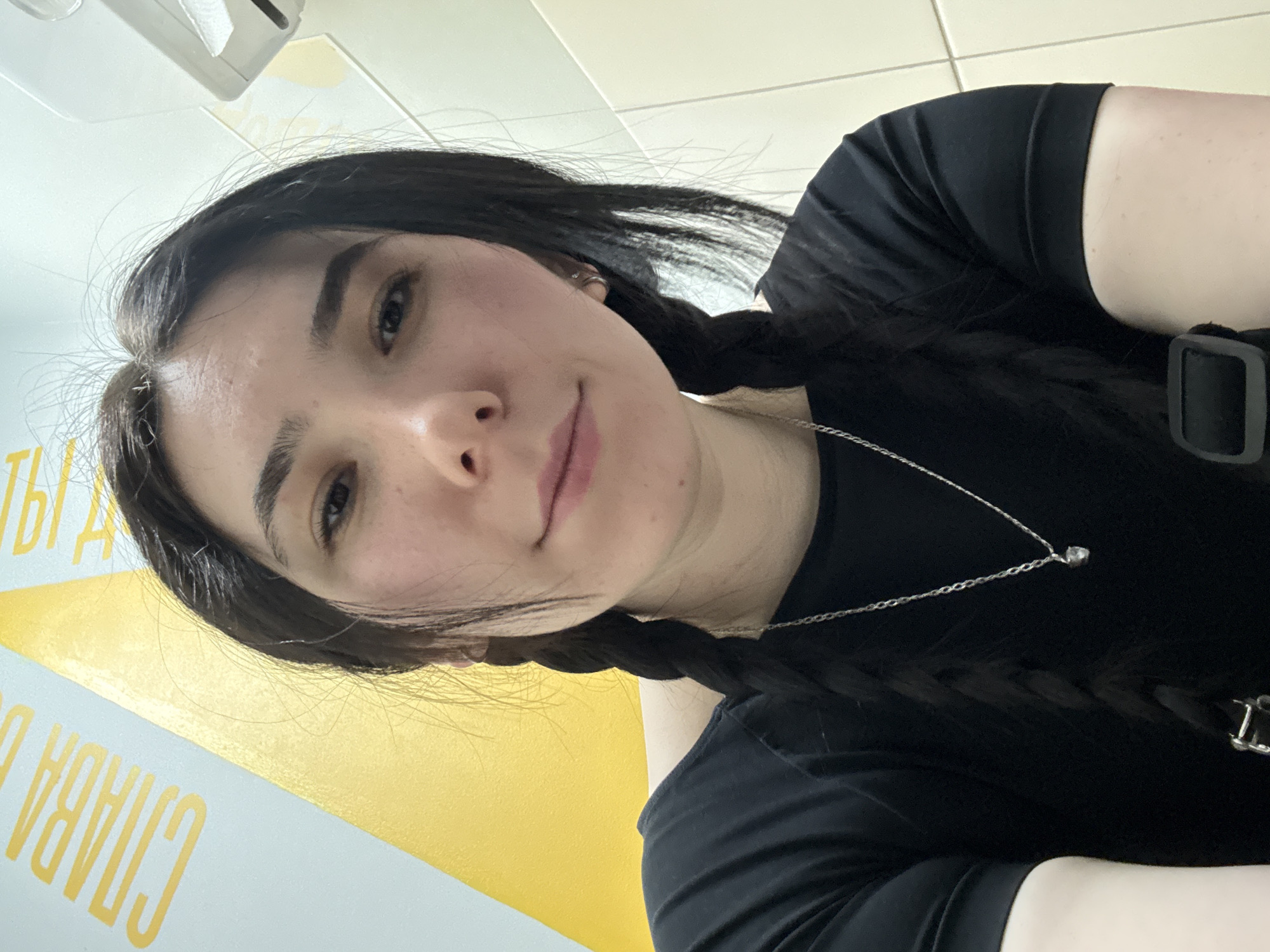 A woman with dark hair braided into two pigtails is taking a selfie. She is wearing a black t-shirt and a silver necklace with a small pendant. The background is a wall with a yellow and white design, featuring the Russian words "Слава труду" (Slava trudu), which translates to "Glory to Labor." This slogan and the surrounding environment suggest the photo was taken in Izhevsk, Russia, where this socialist-era slogan is prevalent.