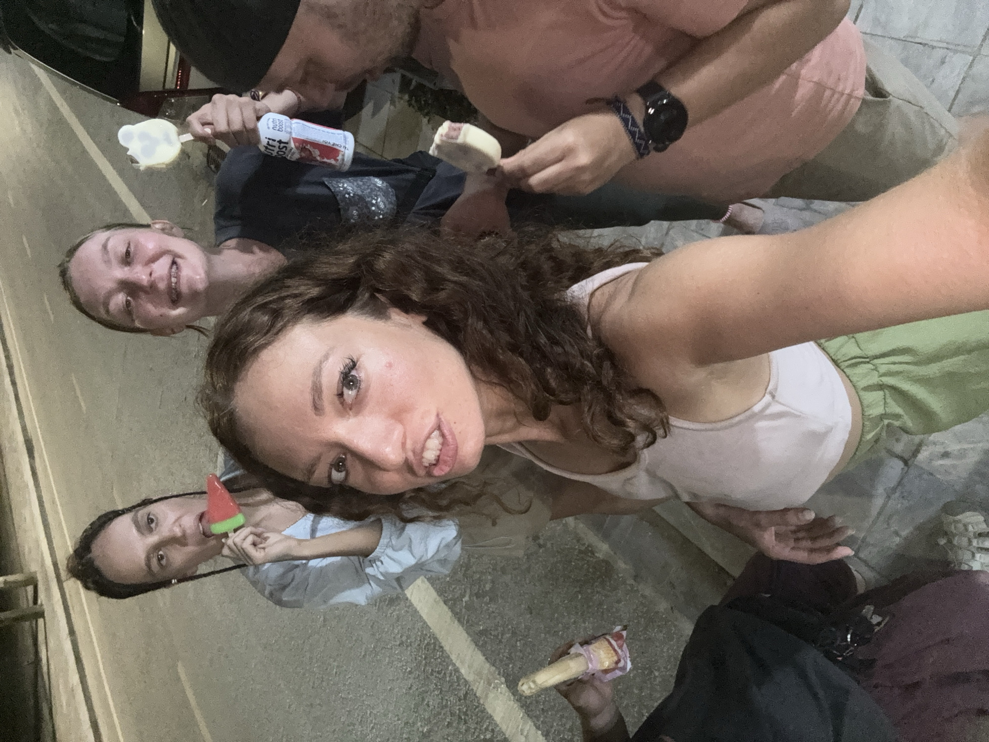 Four individuals are seen in the photo, holding and possibly consuming ice cream in a street setting. One person is holding a watermelon-shaped ice cream. Another individual holds a white ice cream product, while another has a Nutri Boost beverage. The ground surface appears to be tiled in some areas and asphalt in others. It is nighttime. The location is not specified as Yên Minh Commune, Vietnam.