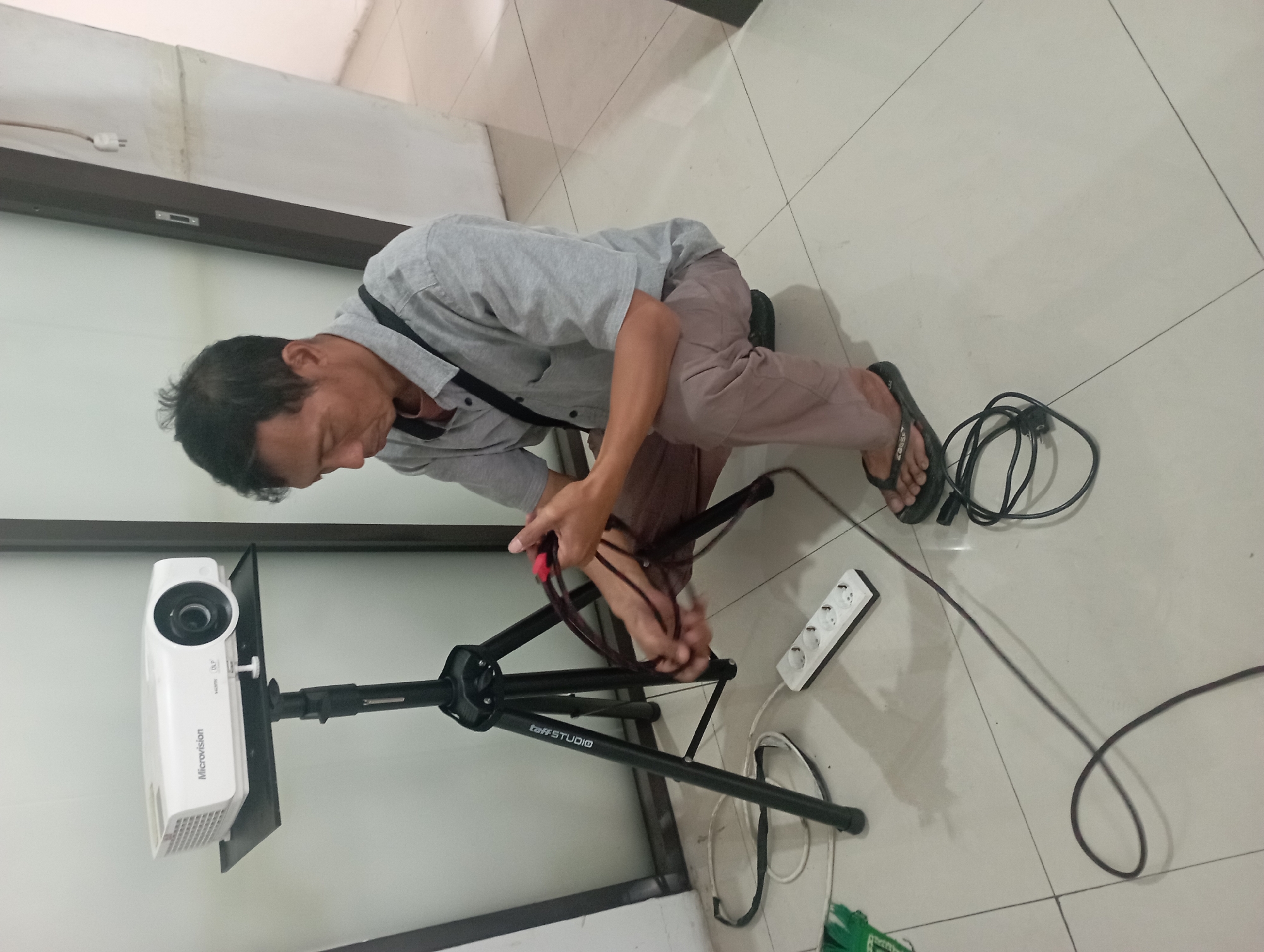 A man is crouching on a tiled floor in front of a projector on a tripod. The man is wearing a grey collared shirt, brown pants, and dark flip-flops. He is holding black cables with red connectors in his hands, appearing to be connecting or disconnecting them. A white power strip with three outlets is visible on the floor, connected to a black cable. The projector, a white Microvision model, is positioned on a black platform atop a black tripod. The environment appears to be an indoor space with a white wall and a doorway in the background. The scene is devoid of any clear indications of the specific city of Tasikmalaya, Indonesia.