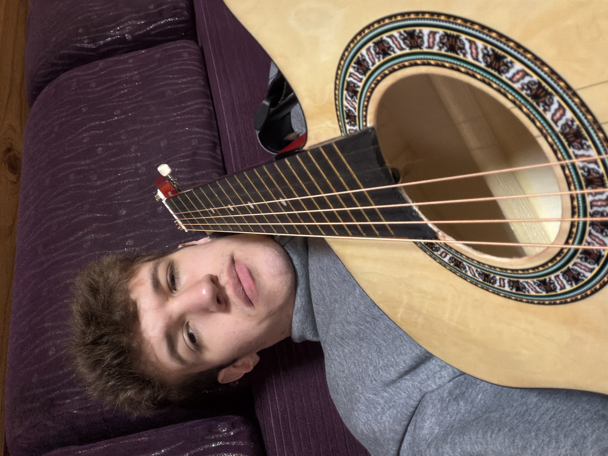 A young male lies on a purple, textured couch, possibly in Прикамский, Russia. He wears a grey turtleneck sweatshirt. An acoustic guitar is positioned over his body, with the neck near his head and the sound hole near his chest. The guitar's soundhole has a decorative rosette. The person is looking upward, and the guitar is oriented in a way that partially obscures his face. The scene appears indoors.