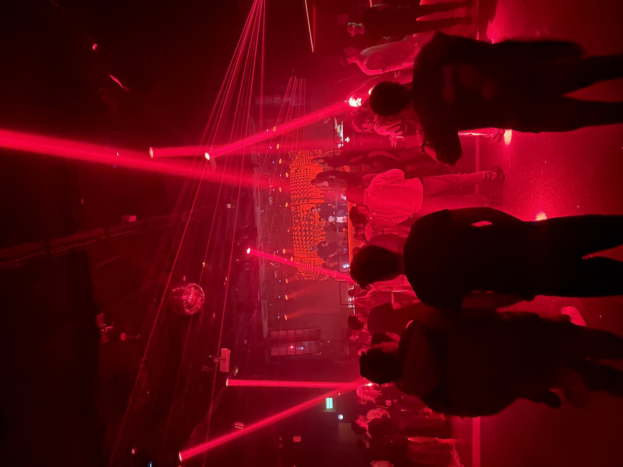 The image shows a crowded nightclub in what is likely Barcelona, Spain, indicated by an external sign. The scene is dimly lit, with a prominent red laser light display illuminating the space. There are numerous people present, mostly facing towards a central platform where a DJ or performer appears to be located. The crowd is dense, obscuring details of individual people. A disco ball is visible in the upper left. The overall atmosphere suggests a lively, active nightlife environment.