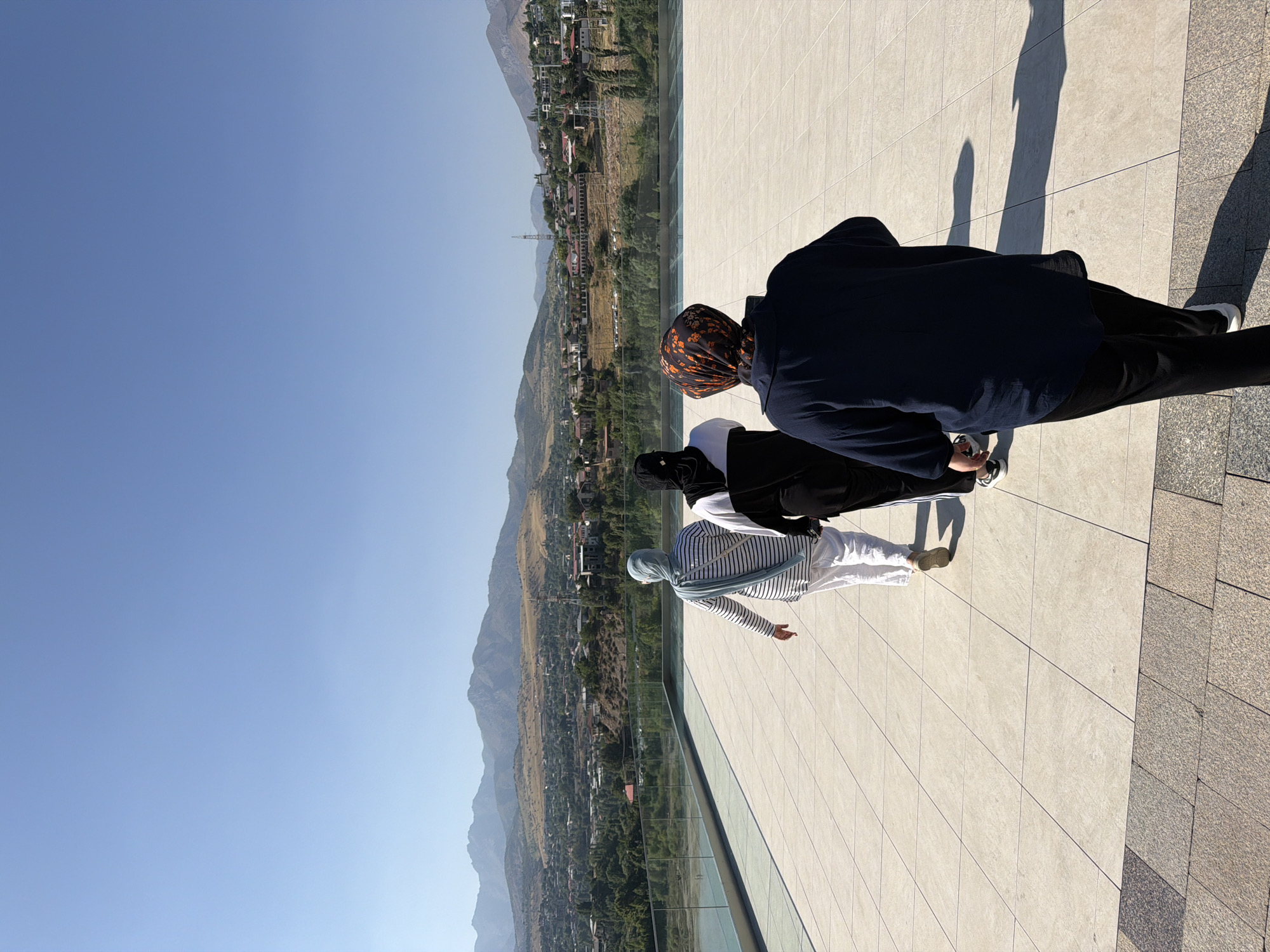 In the city of Chinarkent, Uzbekistan, three individuals are walking away from the camera on a paved outdoor terrace. The terrace is made of light-colored square tiles, with a darker, patterned tile border in the foreground. A modern glass railing runs along the edge of the terrace, overlooking a body of water to the left and a cityscape with mountains in the background.
The person in the foreground is wearing a dark blue oversized top and black pants, with a headscarf featuring an orange and black floral pattern. To their left and slightly ahead is another individual in a black and white tracksuit, also wearing a black headscarf that covers most of their face. Furthest left and ahead is a person in a white and blue striped long-sleeved shirt and white pants, with a light blue headscarf. All three appear to be walking in the same direction, away from the viewer. The sky is clear and blue, and the sun is casting long shadows on the terrace. The city of Chinarkent is visible below, nestled in a valley with buildings and vegetation, with rolling, dry hills and mountains forming the backdrop.