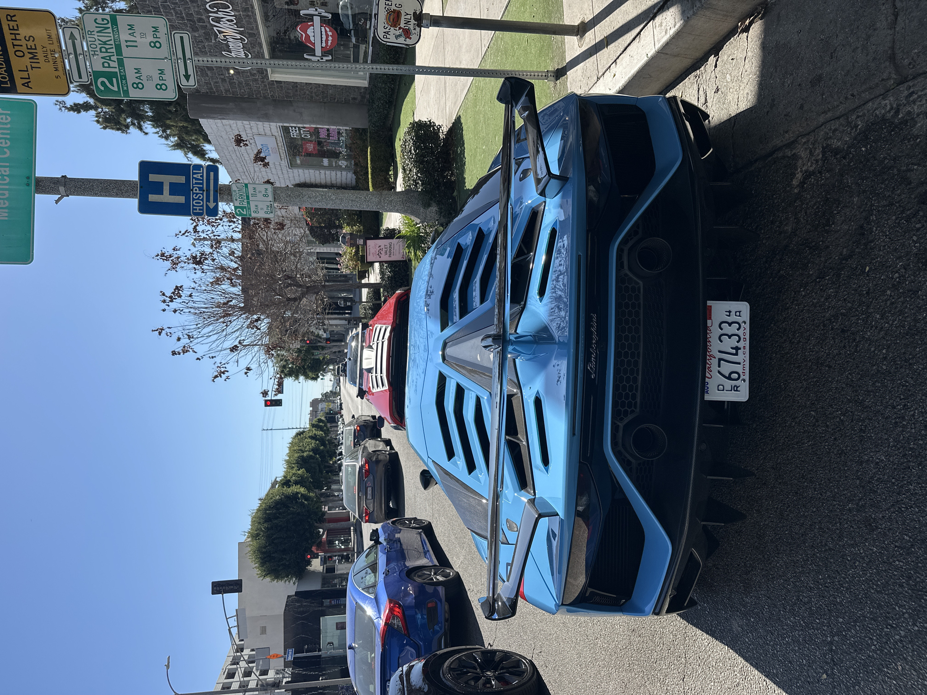 The image captures a vibrant street scene on a clear, sunny day, with a luxurious blue sports car prominently featured in the foreground. The photo is rotated 90 degrees counter-clockwise, making the street appear to run vertically from the bottom of the frame to the top.
In the immediate foreground, a striking bright blue Lamborghini sports car is parked on the side of the road, facing away from the viewer. Its aggressive rear design is clearly visible, featuring a large rear spoiler, intricate vents on the engine cover, and dual large, circular exhaust pipes integrated into a dark diffuser. The word "Lamborghini" is inscribed in cursive script on the rear of the car, above the grille. A California license plate is affixed to the back, reading "AUG / California / L 67433 / dmv.ca.gov". Part of a wheel from another vehicle, possibly a dark blue sedan, is visible in the bottom left corner, parked behind the Lamborghini.
Further down the street, several other cars are either parked or moving slowly in traffic. A red sports car, possibly a Ferrari, is visible in the middle distance, followed by a line of other passenger vehicles of various makes and colors. The street is lined with mature trees, some with bare branches and others with green foliage.
To the right of the street, there's a paved sidewalk and a curb, beyond which are commercial buildings. One storefront displays a prominent, stylized red lips logo with "Luxury Wash" text partially visible. Another building has a sign that reads "FREE CITY" and additional text. A "VALET PARKING" sign, set on an easel, is also visible on the sidewalk. Nearby, a small sign reads "PASSERBY ONLY / 5 MIN. G".
Multiple street signs are clearly visible overhead, attached to poles. These include:
* A green rectangular sign with an arrow pointing right, stating "2 HOUR PARKING / MON-SAT / 8 AM TO 8 PM / SUNDAY / 11 AM TO 8 PM".
* A yellow rectangular sign with an arrow pointing left, indicating "LOADING / ALL OTHER TIMES / DAILY / 5 MINUTE LIMIT / PROPERTY OF CITY OF LOS ANGELES".
* A blue square sign with a white "H" and "HOSPITAL" below it, accompanied by an arrow pointing right.
* A smaller, partially visible green "2 HOUR PARKING" sign is further down the street, likely with similar parking regulations.
The sky above is a clear, bright blue, indicating good weather. Shadows cast by poles and buildings suggest it is either mid-morning or early afternoon. A traffic light showing a red signal can be seen in the distance, further down the street. The overall setting appears to be a busy commercial street in a Californian city, likely Los Angeles, given the text on one of the signs.