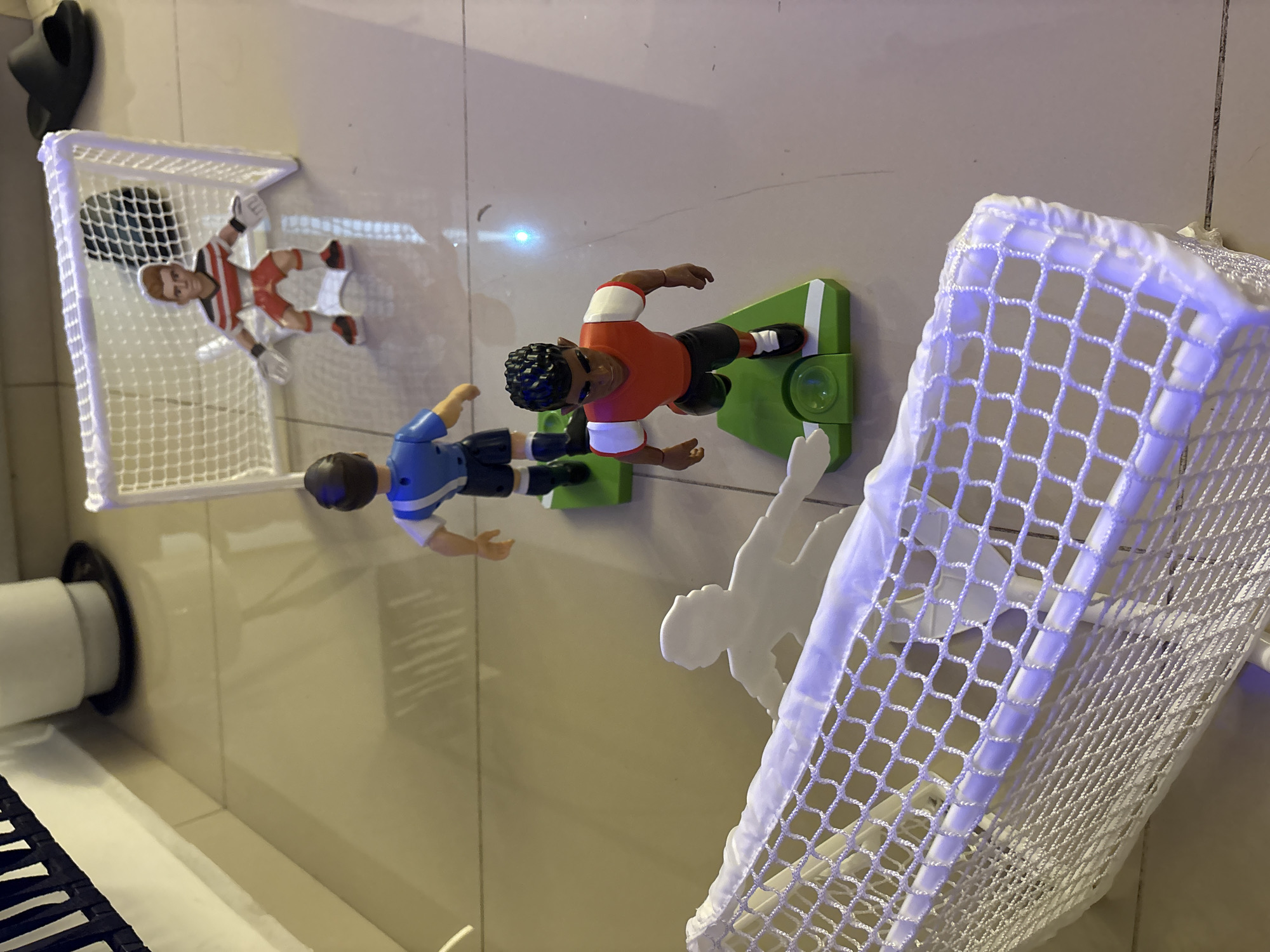 The image depicts a tabletop foosball setup on a light-colored, reflective surface, likely a table or floor. Two foosball goals, constructed from white mesh netting and plastic frames, are positioned on opposite ends. Four foosball players are visible: one wearing a red and white goalie uniform within the left goal, one wearing a blue jersey and black shorts, and another wearing an orange jersey and black shorts facing the right goal. A flat white soccer player figure is positioned near the right goal. The foosball players are connected to green bases with knobs. Additional details include a black slide, and a black and white striped object on the left side of the image. No geographic information is discernible from the image.