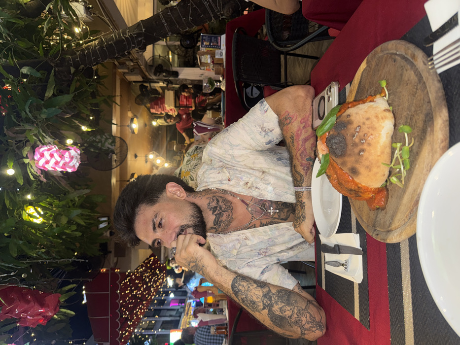 An evening scene depicts a male individual seated at an outdoor restaurant table in Pattaya City, Thailand. The man, appearing to be in his late 20s to mid-30s, possesses dark hair with faded sides, a trimmed beard, and extensive tattoos covering his neck, both forearms, and visible chest. He wears a light-colored, patterned short-sleeved collared shirt, multiple silver necklaces (one with a cross pendant), a silver bracelet, and a ring on his right hand. He is looking off to his left, with his right hand near his chin.
The table is draped with a red tablecloth and features a dark placemat with horizontal stripes. On the placemat rests a white plate, a fork, and a knife. Centered on a round wooden board in front of the man is a partially baked dough dish, resembling a calzone, topped with red sauce and green leaves. An iPhone in a clear case is on the table to the man's right. Another white plate is visible in the foreground.
The environment is an open-air dining area illuminated by numerous small string lights strung through overhead tree branches. Decorative lanterns, one red and one pink-and-white patterned, hang from these branches. In the background, other patrons are seated, and restaurant staff wearing red shirts are visible further back. The establishment's interior shows hanging lights and wall-mounted fans. Beyond the dining area, blurred lights indicate a busy urban street at night. A large tree trunk, also adorned with string lights, is on the right side of the frame.