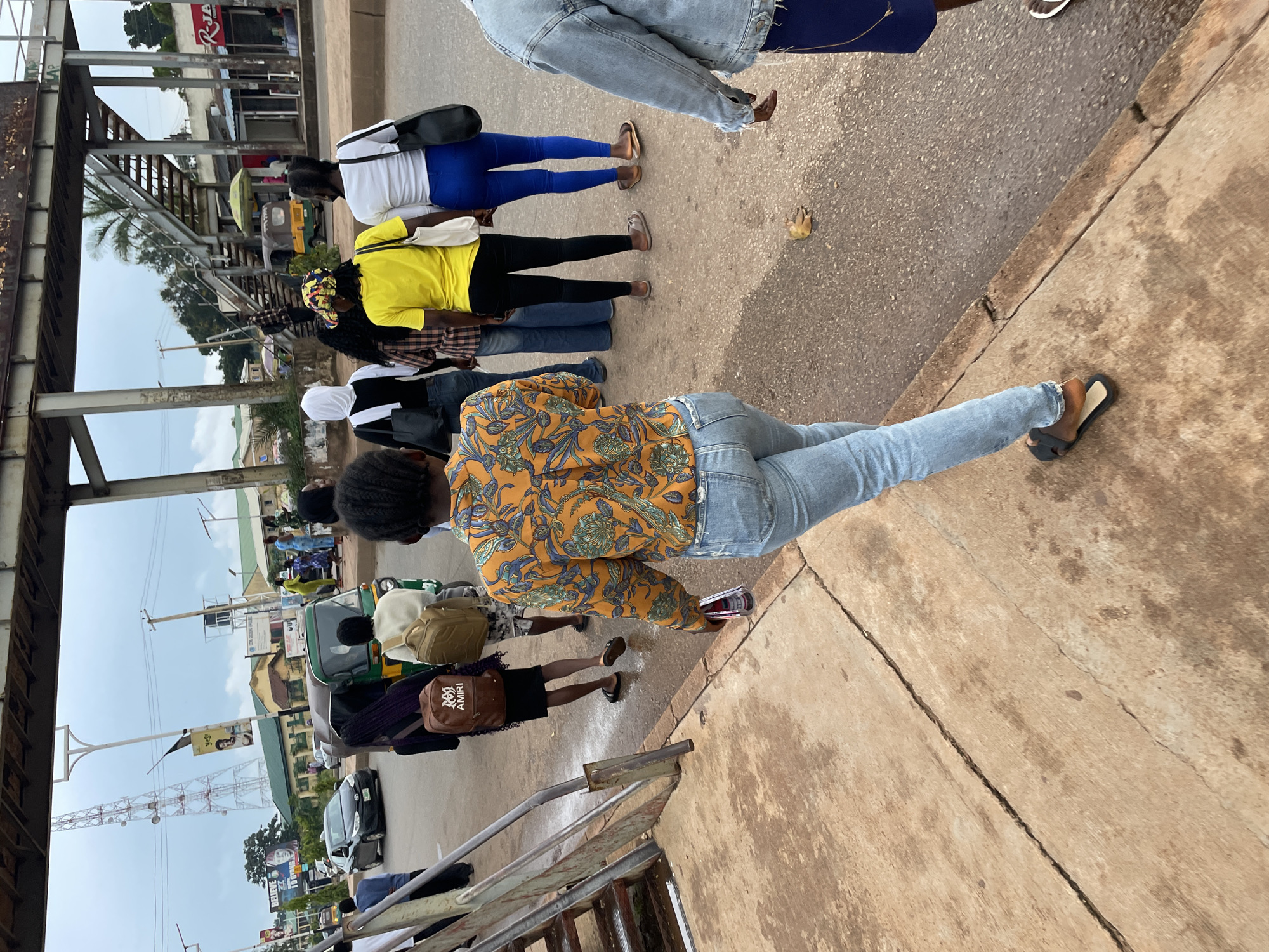 The image depicts a street scene in Jos, Nigeria, under an overcast sky. In the foreground, a young woman with braided hair, wearing a patterned orange long-sleeved shirt and ripped light blue jeans, walks away from the camera on a cracked concrete sidewalk. She is holding a clear cylindrical object in her right hand. Behind her, a group of people are walking on the asphalt road. One woman in the middle wears a yellow t-shirt and black pants, carrying a white tote bag. To her left, another woman in a white headscarf and black top carries a black tote bag. Further back, a person with a backpack is visible. A green and yellow auto-rickshaw is also present on the road, alongside other vehicles and a pedestrian bridge in the background. Buildings and a telecommunications tower are visible on the horizon. The overall impression is of an urban environment with daily activity.