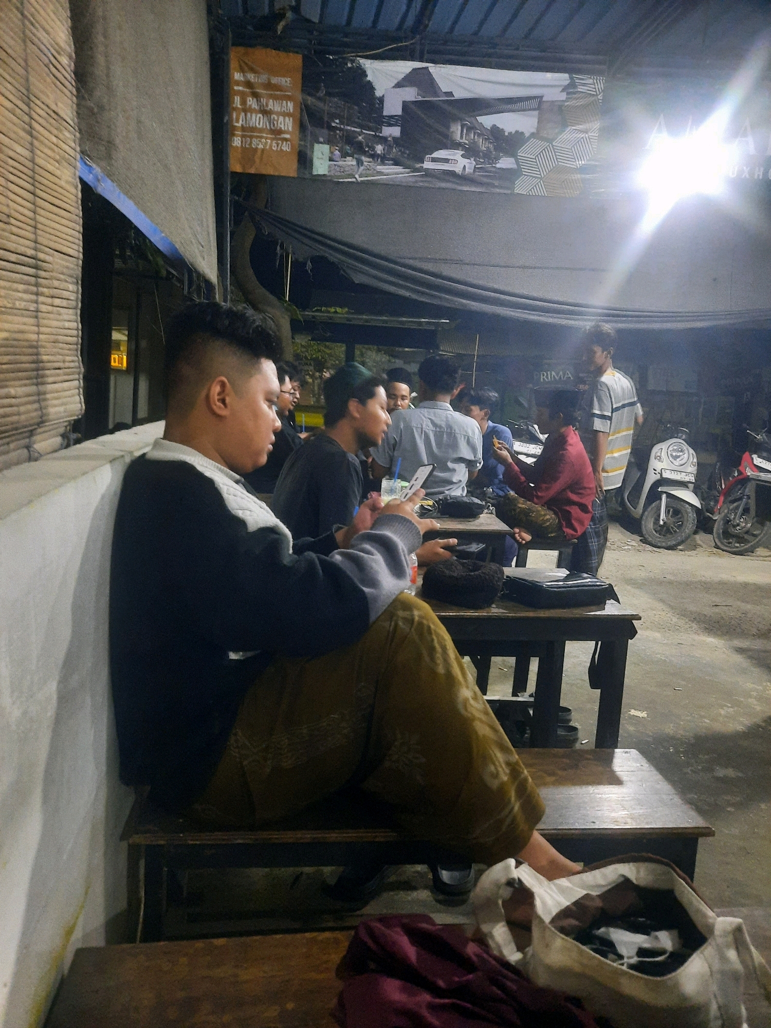 In the city of Pucangtelu, Indonesia, a group of men are gathered at an outdoor establishment at night. The scene is illuminated by artificial lighting. A man in the foreground, facing right, wears a dark blue and grey sweater and olive-colored patterned pants. He is seated on a wooden bench, with his legs crossed. To his left, a white wall with a bamboo-like texture is visible, and a blue tarp hangs above. A sign on the wall displays "MARKETING OFFICE JL. PAHLAWAN LAMONGAN 0812 8557 6740."
In the background, several other men are seated at wooden tables. One man in a red shirt is holding something in his hand. Another man is wearing a grey and white striped shirt. Two scooters are parked nearby, one white and one red. The ground appears to be concrete. A large banner hangs above, displaying architectural imagery and text, including what appears to be "ALANA LUXHO..." and "PRIMA." A white canvas bag filled with items sits on the floor in the foreground, next to a folded burgundy cloth.
