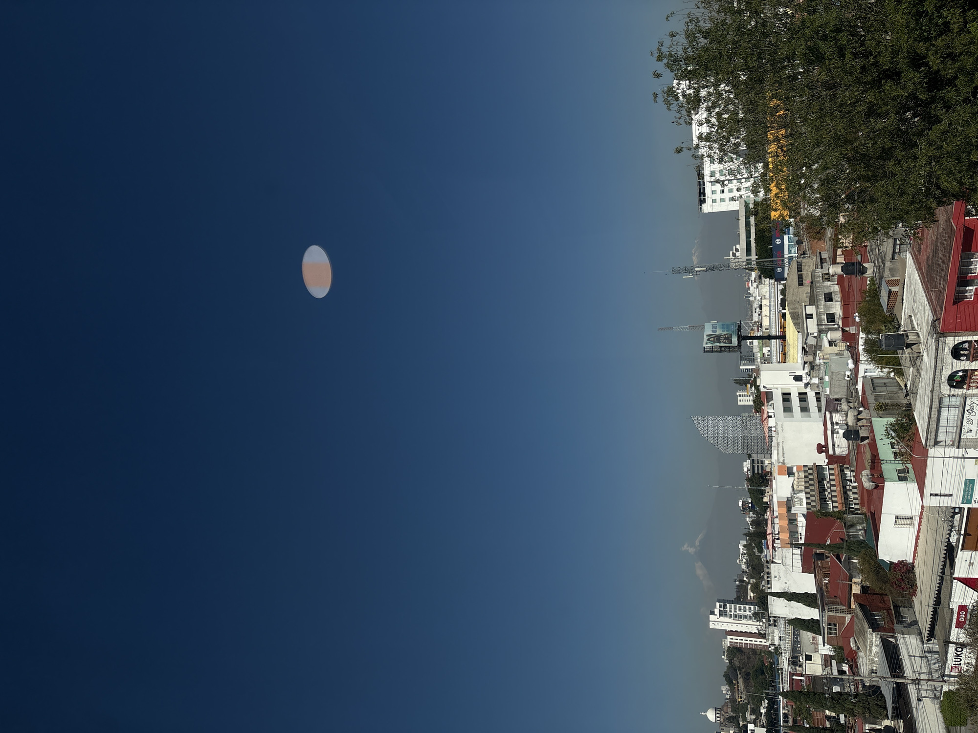 The image depicts an upside-down aerial view of Puebla, Mexico. A bright orb, possibly a reflection or lens flare, is visible in the upper left against a deep blue sky. The cityscape, occupying the right side of the image, consists of densely packed buildings with predominantly red tile roofs, interspersed with white and grey structures. Lush green trees are seen at the edge of the frame. Towers and antennas are visible among the buildings. Street signage is barely legible on the lower right. The sky transitions from a darker blue to a lighter, hazy tone towards the horizon.