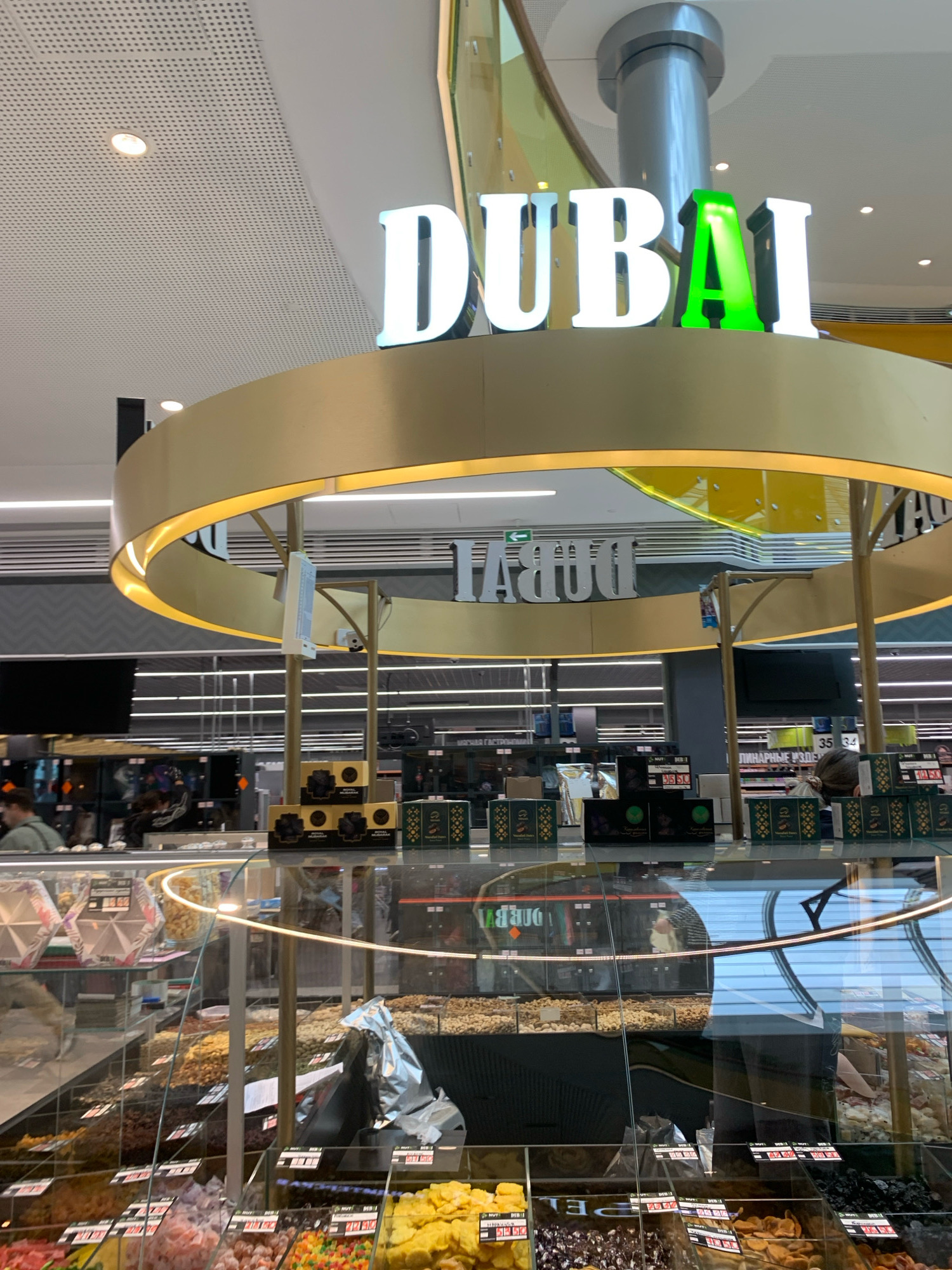 The image shows a store interior with the word "DUBAI" displayed in large, illuminated letters on an overhead structure. Below the sign, a circular display counter features various dried fruits, nuts, and sweets in transparent bins. The store appears to be a market or specialty food shop. The lighting is bright, and reflections from the ceiling are visible. Several price tags are present on the bins. The overall impression is of a well-stocked retail environment. The city of Minsk, Belarus, is mentioned as the location of this establishment.